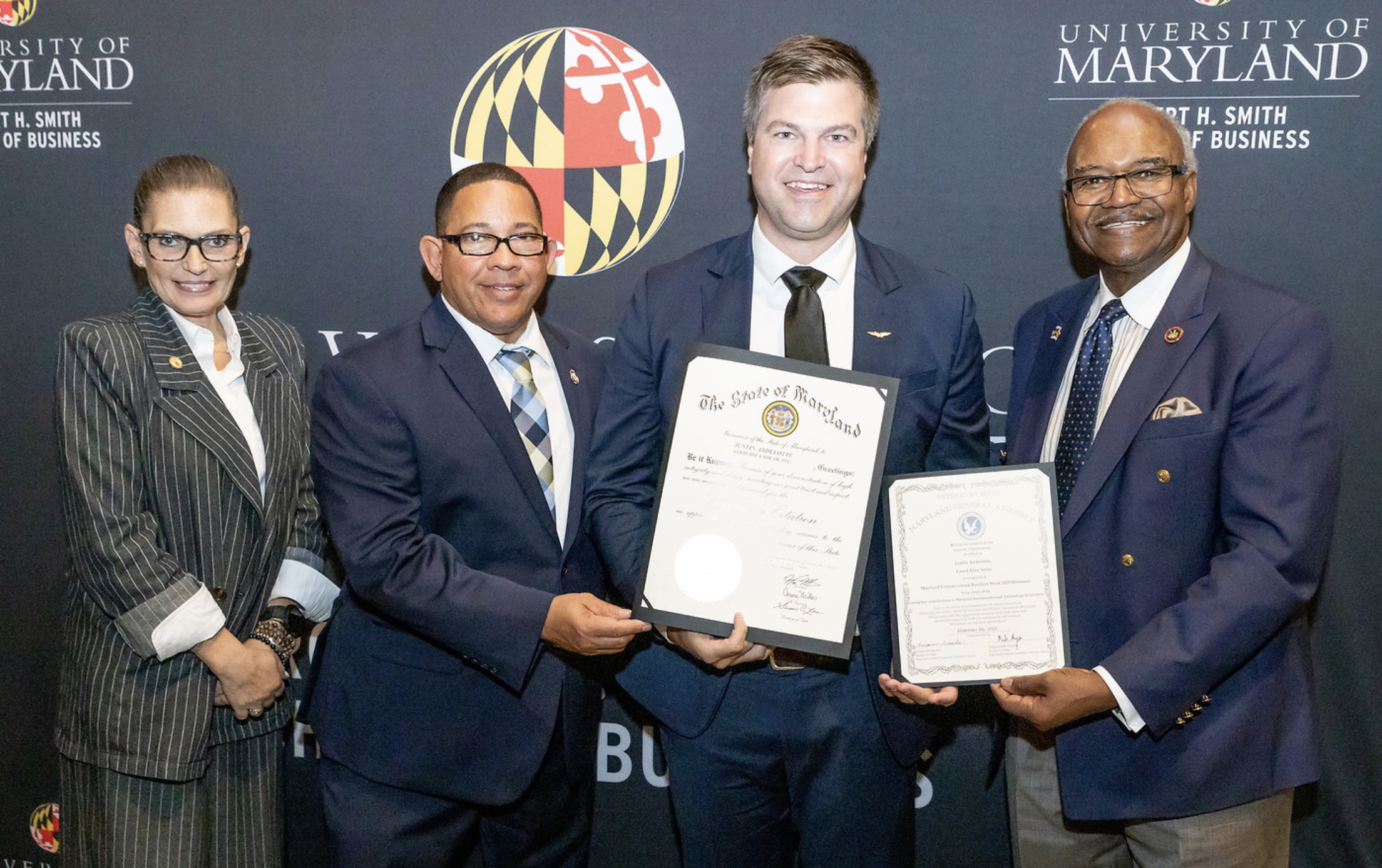 A group of five people in business attire standing in front of a Maryland-themed backdrop, holding certificates and smiling at the camera.