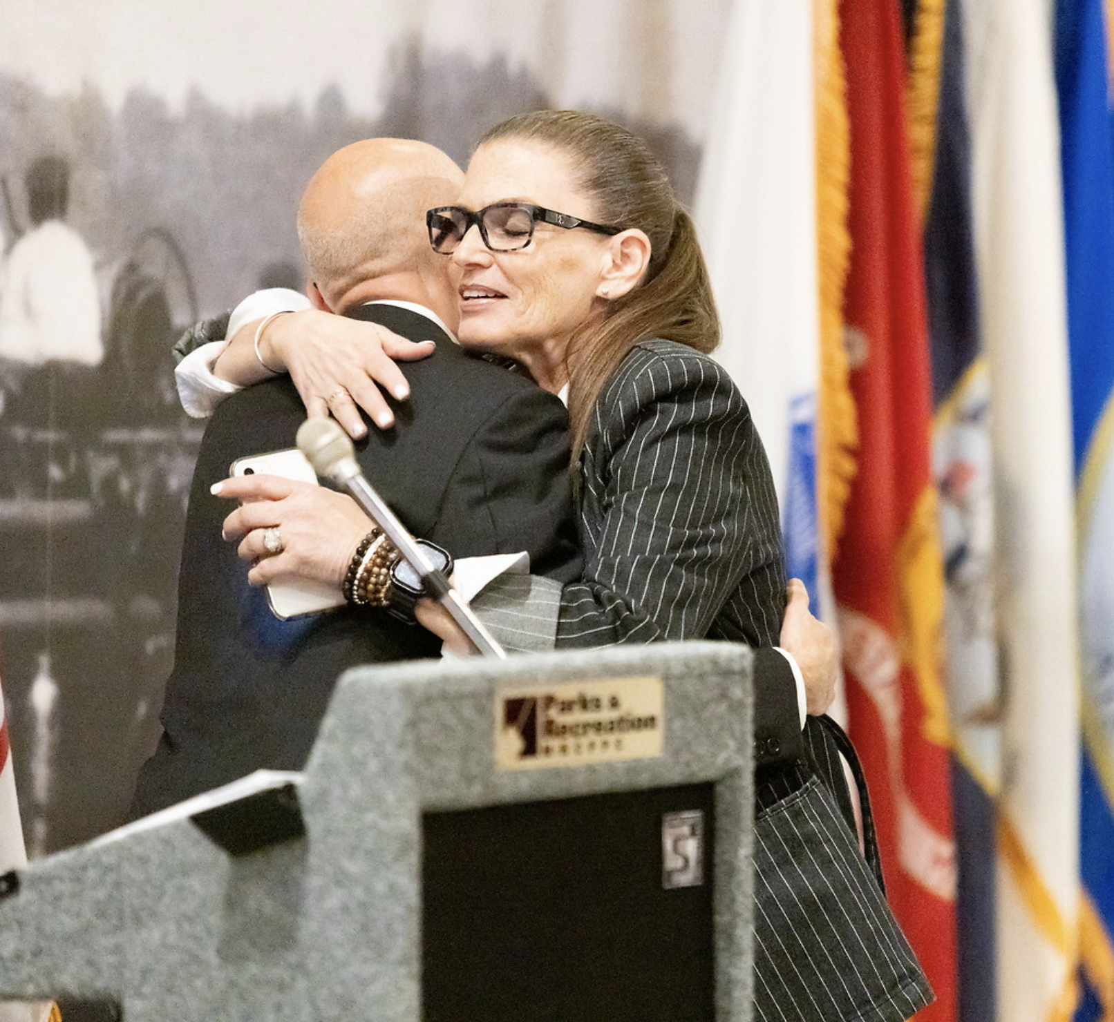 Two women hugging at a formal indoor event, with flags in the background and a podium labeled 'Texas Association of Community Colleges' in the foreground.