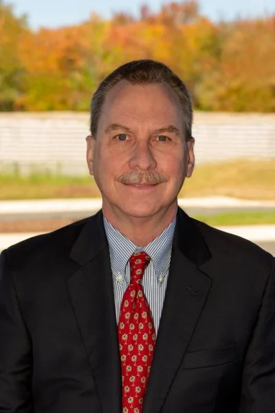 A man in a black suit, blue striped shirt, and red patterned tie, sitting outdoors with autumn trees and a white fence in the background.