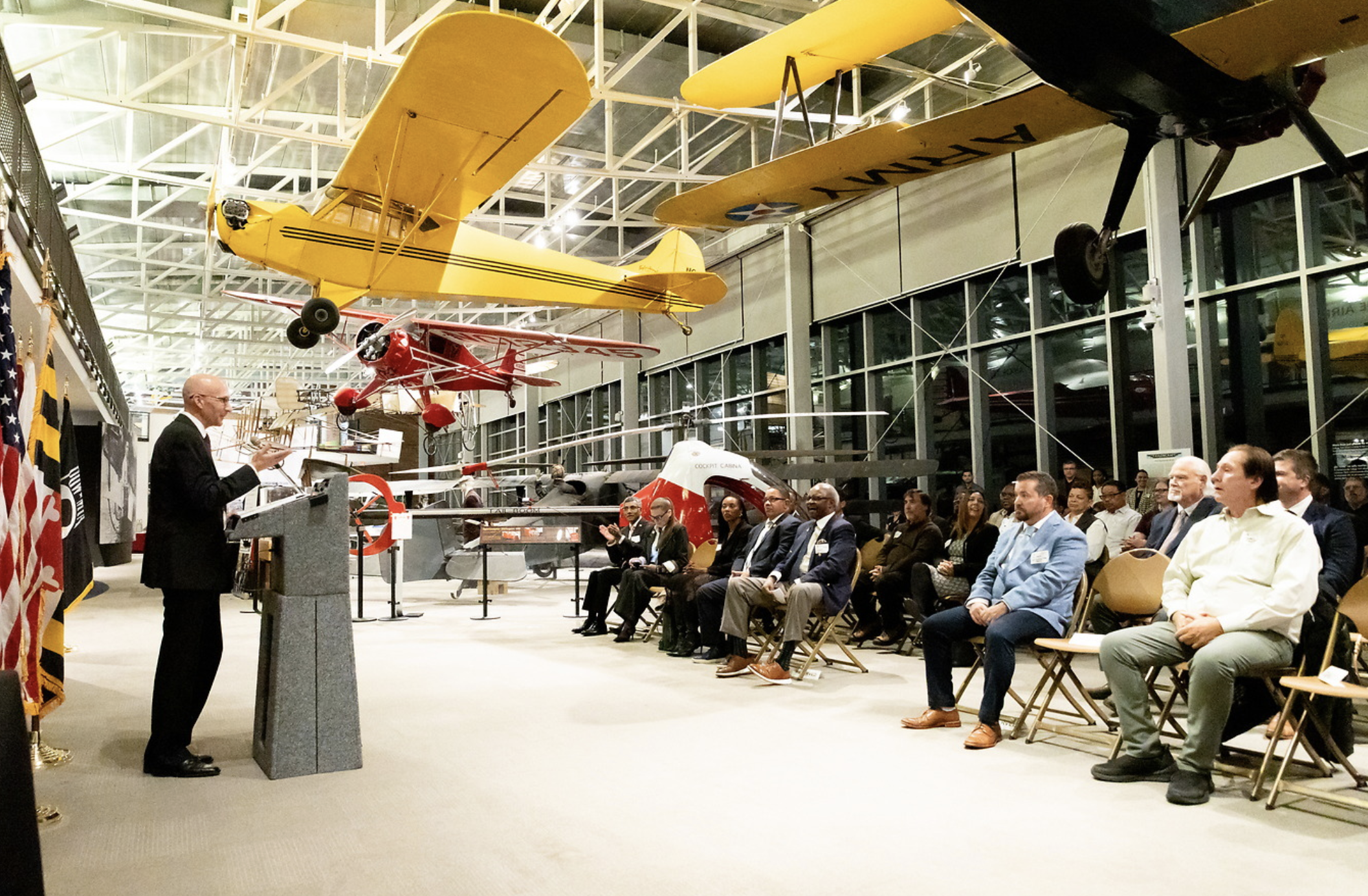 An indoor aviation exhibit with several small vintage airplanes hanging from the ceiling; a man in a suit speaking at a podium; an audience seated and listening; large glass windows in the background.