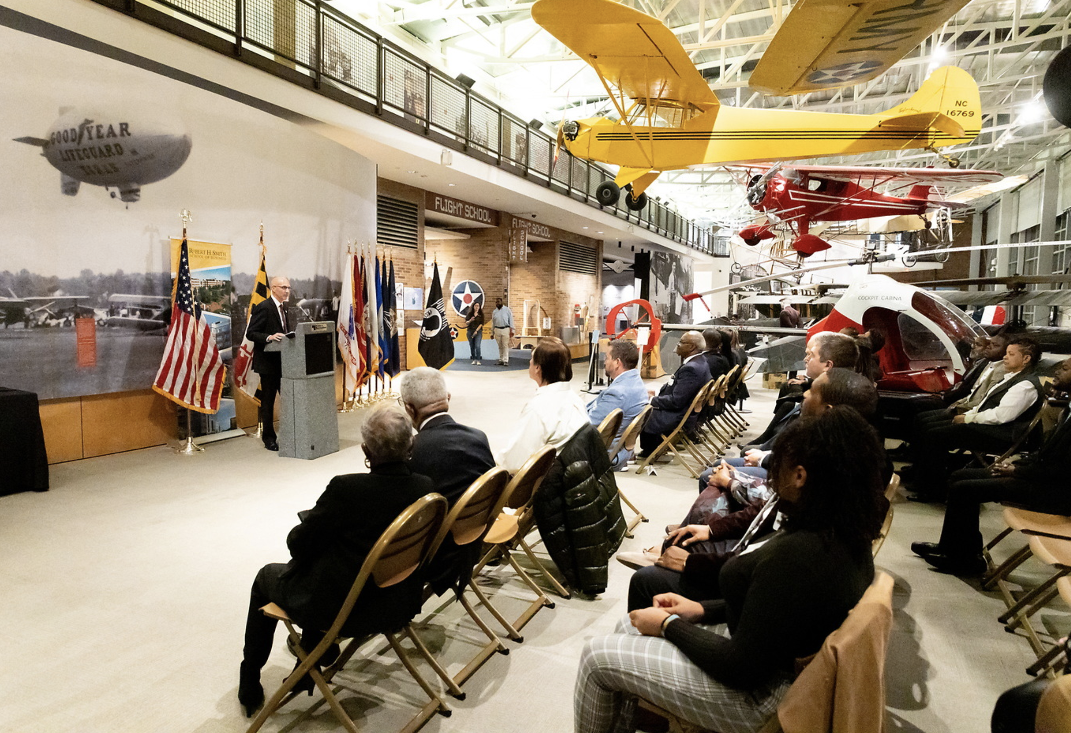 Indoor space with vintage aircraft hanging from the ceiling and seated audience attending a presentation or ceremony, with flags and a speaker at a podium.