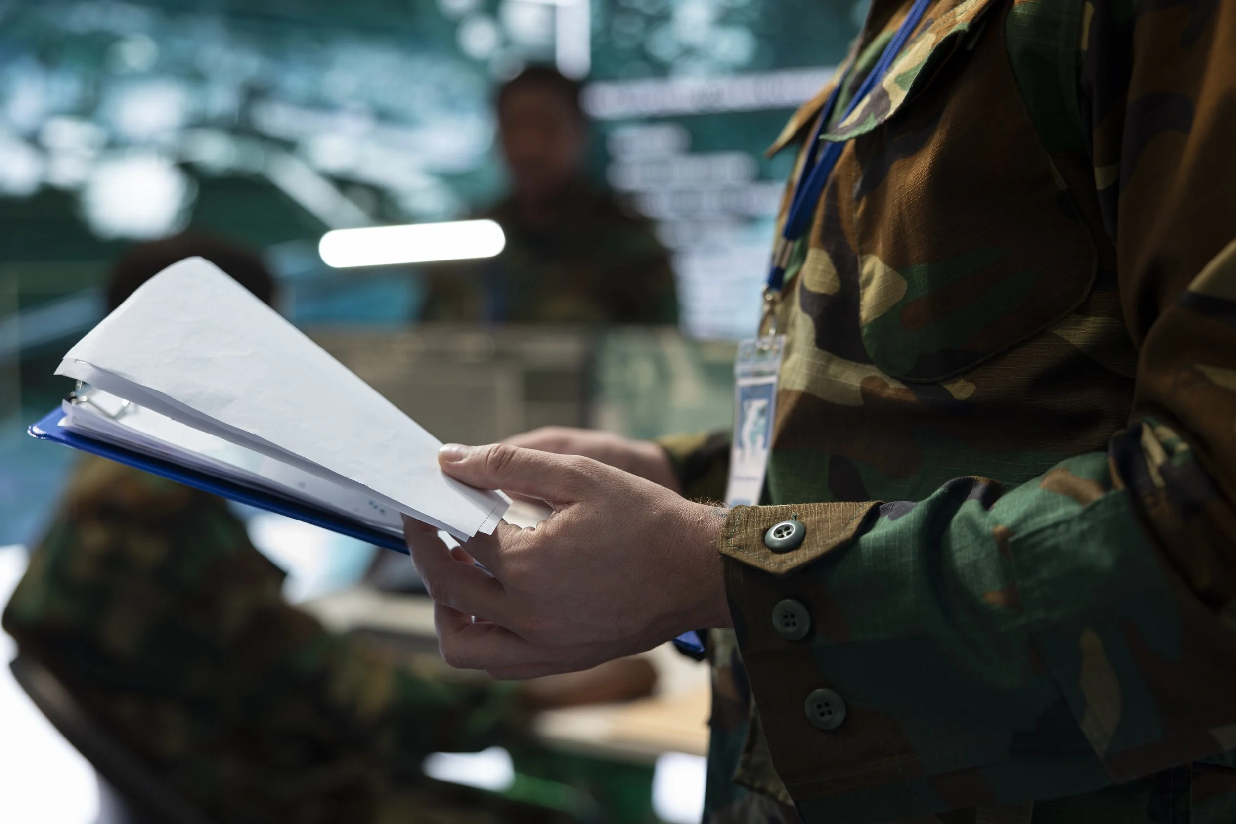 Person in military uniform holding a clipboard and papers, with others in the background.