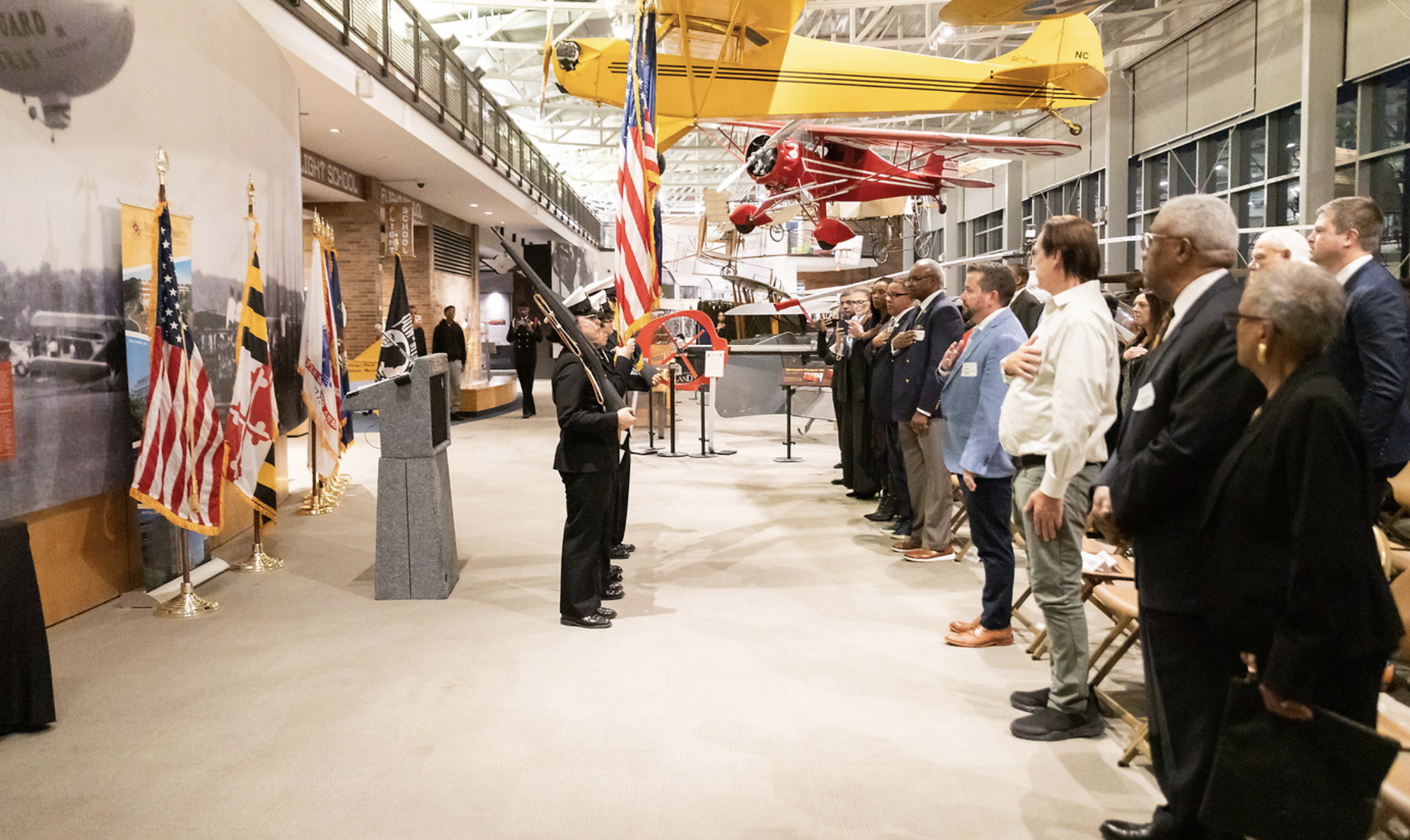 A group of people standing in a line with their hands over their hearts, facing a woman in a military uniform saluting, inside a museum with vintage airplanes hanging from the ceiling and flags along the wall.