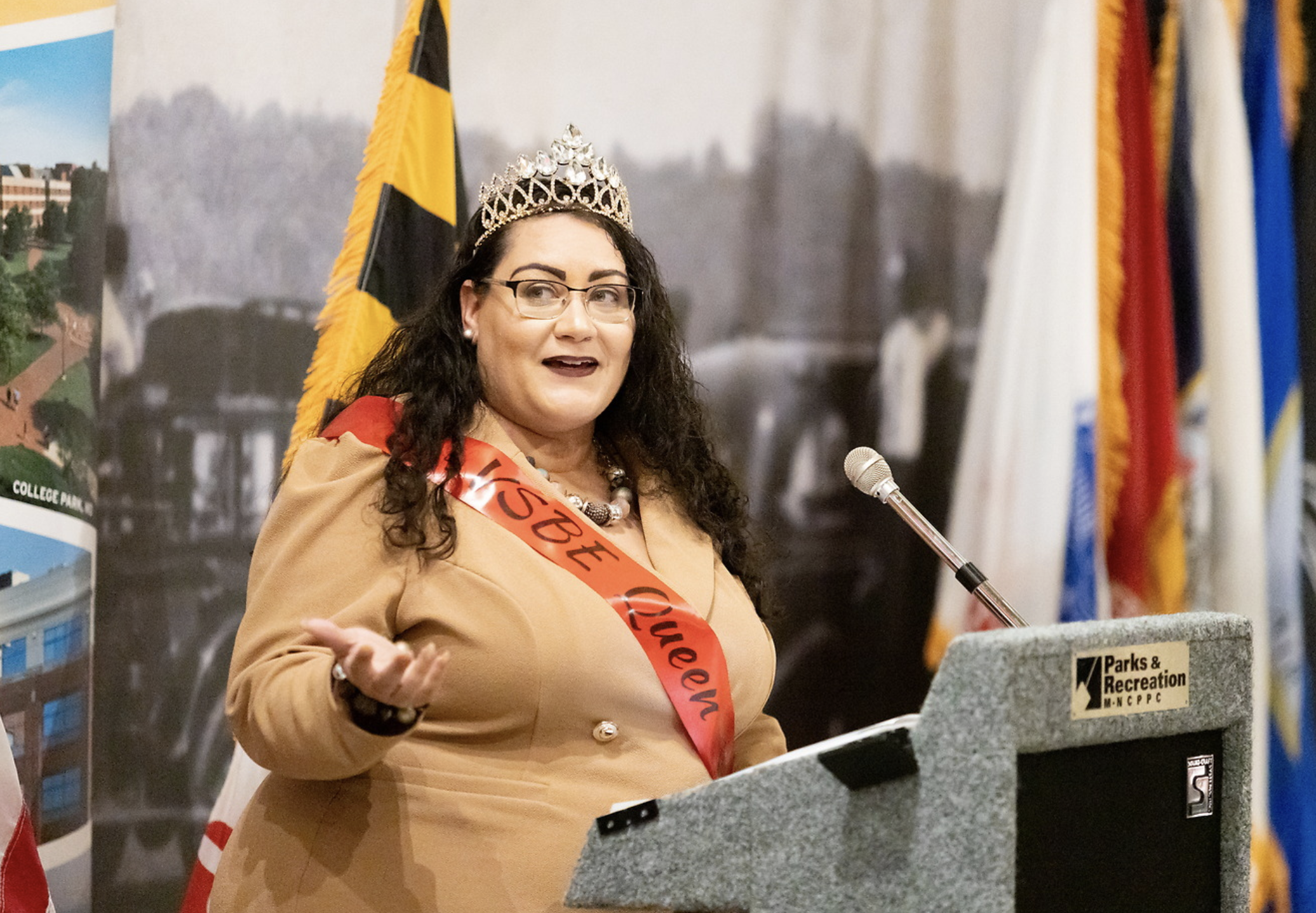 A woman with dark curly hair, wearing glasses, a tiara, a beige blazer, and an orange sash, speaking at a podium with flags and banners in the background.
