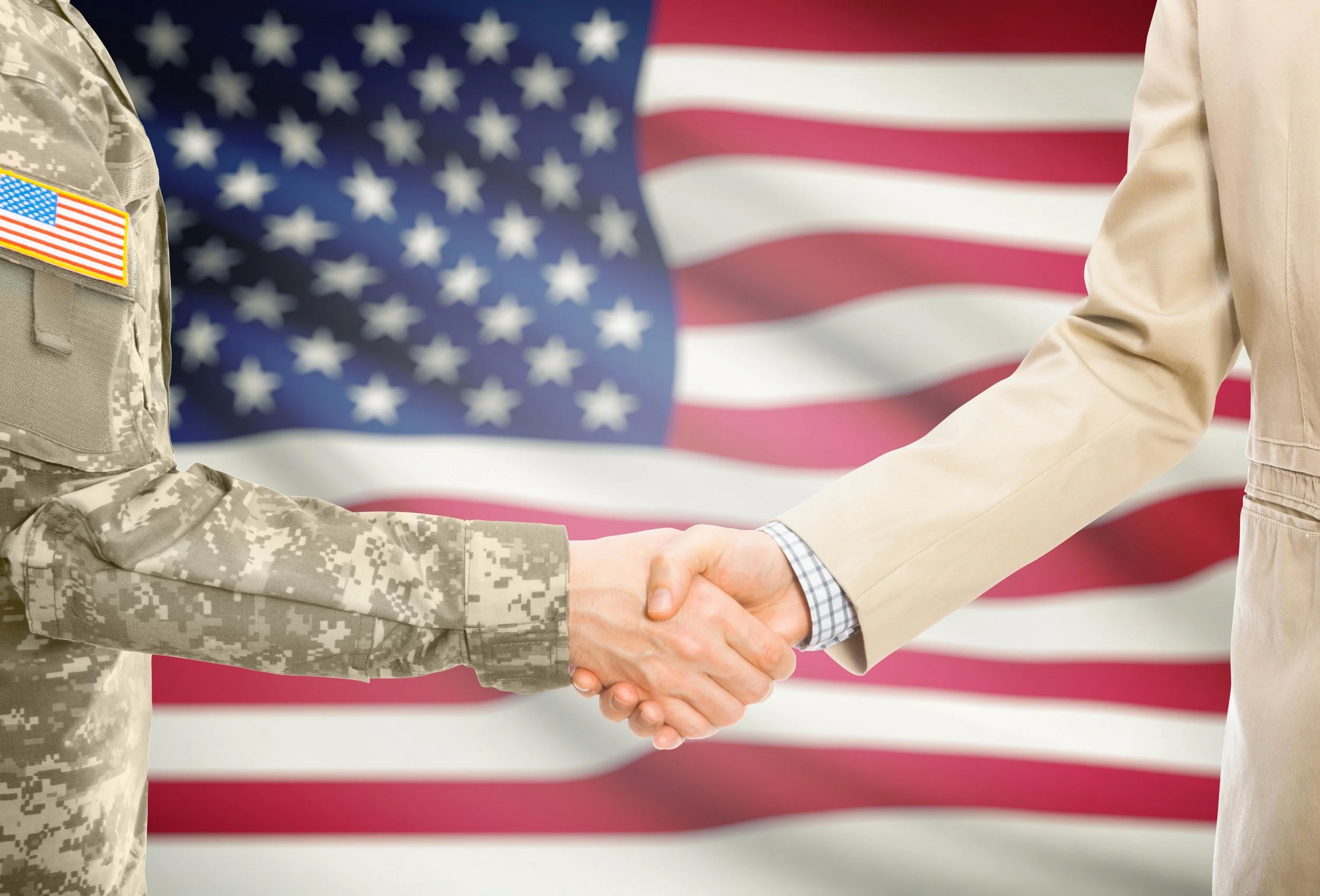 A soldier in camouflage uniform shaking hands with a person in a beige suit in front of an American flag.