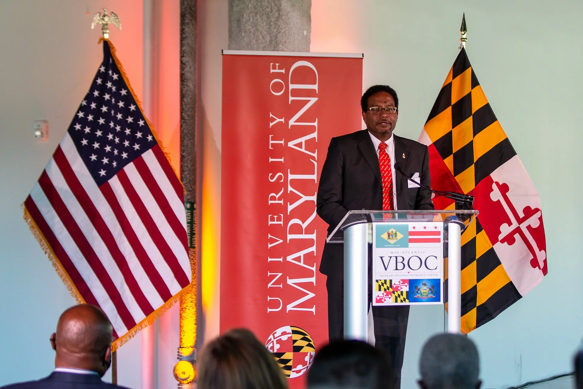 A man in a dark suit, white shirt, red patterned tie, and glasses is standing at a podium, delivering a speech. Behind him are the flags of the United States and Maryland, and a red banner with white text reading 'University of Maryland.' The podium displays a logo for the Veterans Business Outreach Center (VBOC), featuring small flags of Maryland and the United States.
