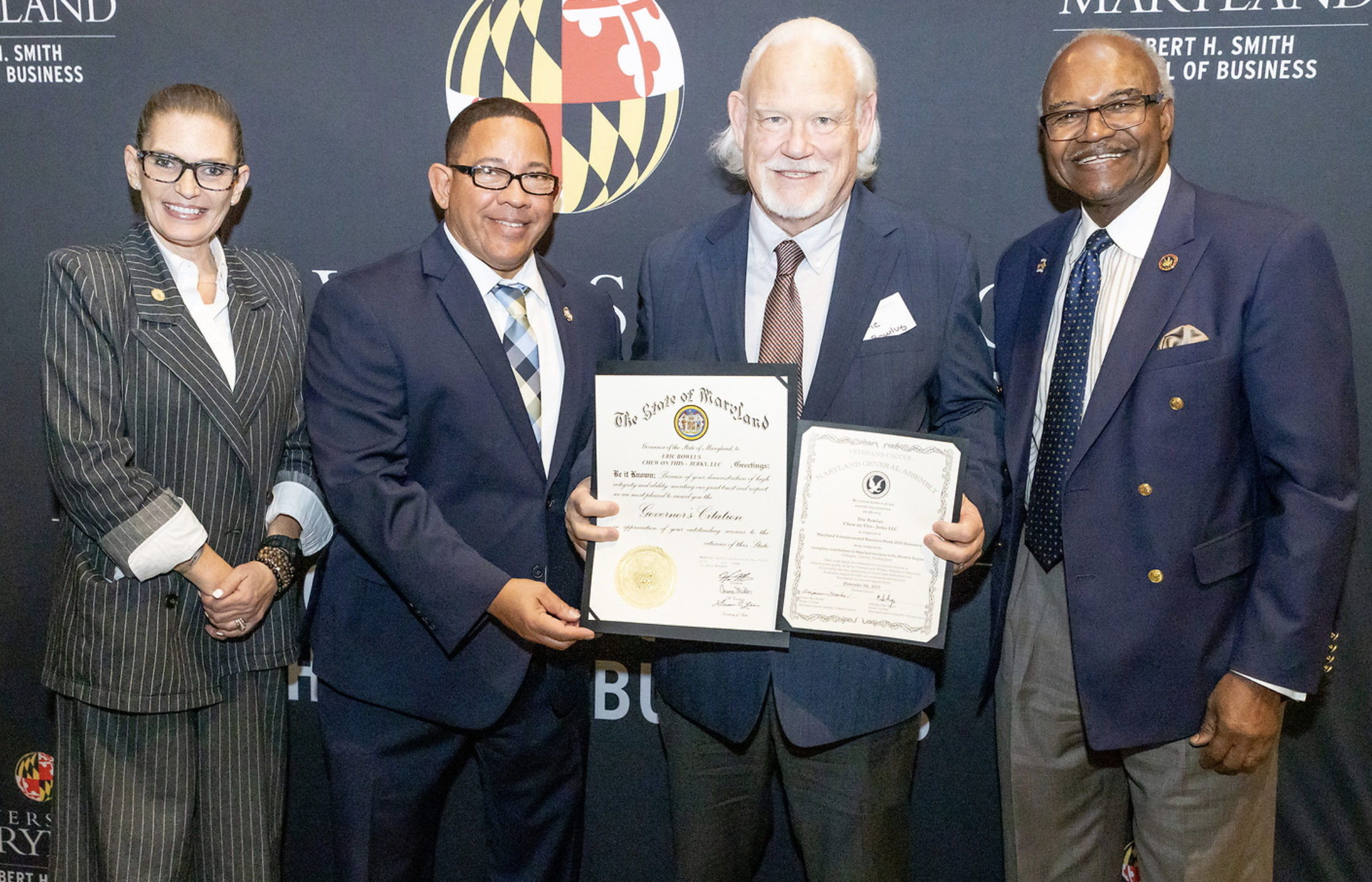 Four professionally dressed individuals standing together at an award or certificate presentation event, with a dark background featuring Maryland-related symbols and texts.