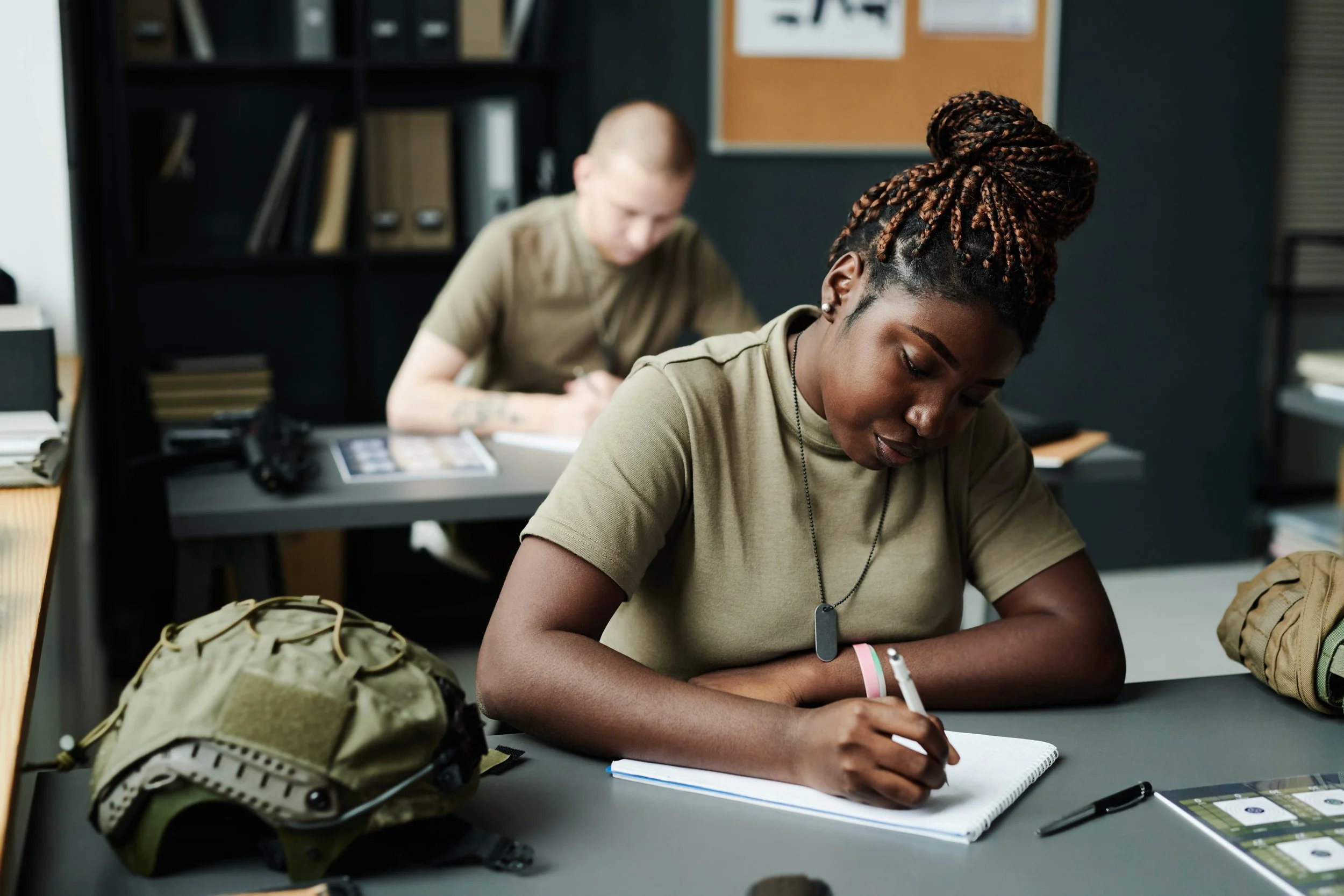Two soldiers, one woman and one man, in a classroom setting. The woman with braided hair is writing in a notebook, and the man is also working at his desk in the background.
