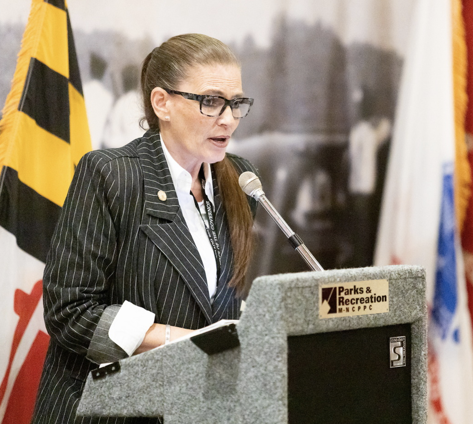 Woman speaking at a podium with a Parks & Recreation sign, behind flags and a blurred background.