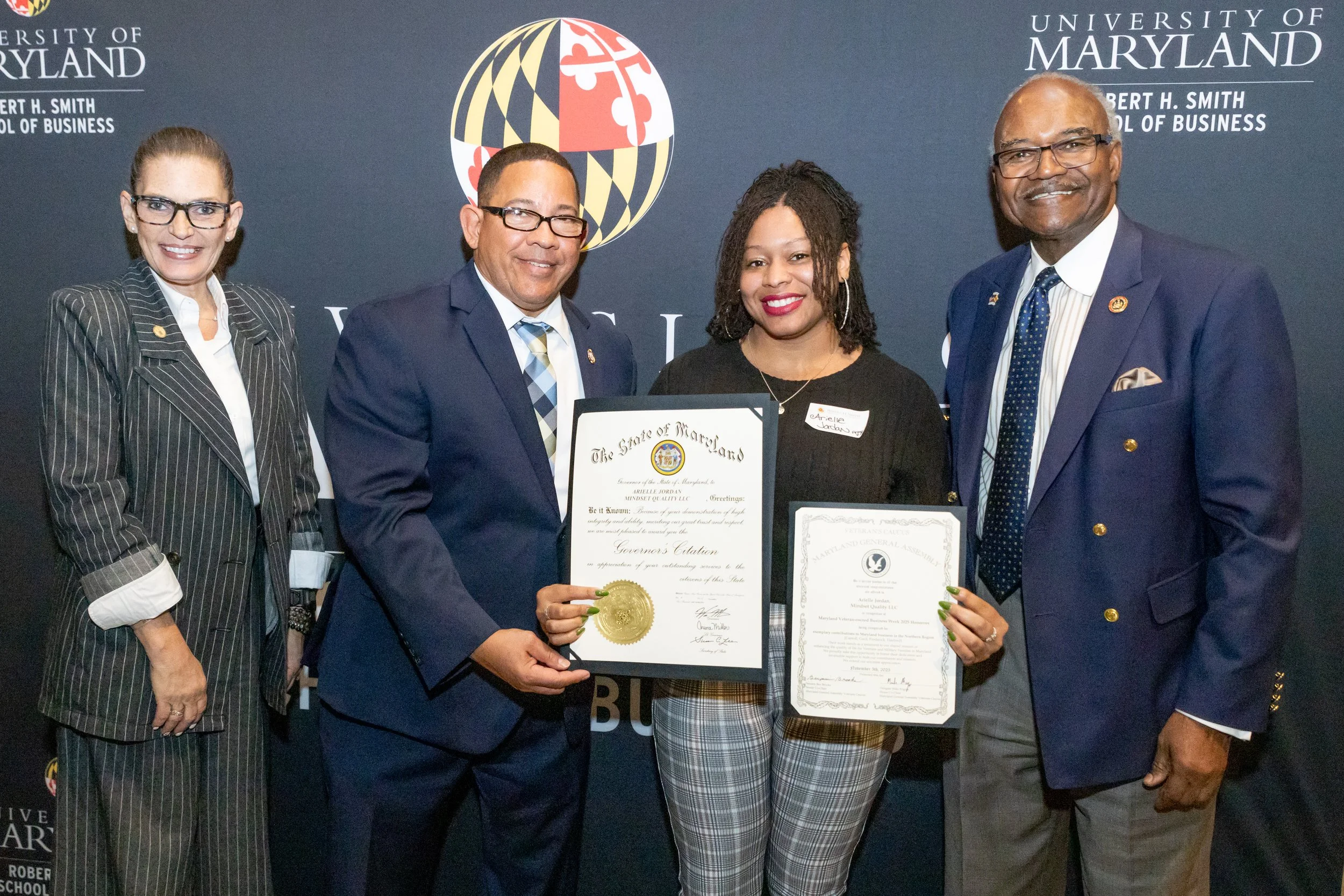 Group of five diverse adults standing together at a graduation or award ceremony, holding certificates and medals, in front of a dark backdrop with university and Maryland state logos.
