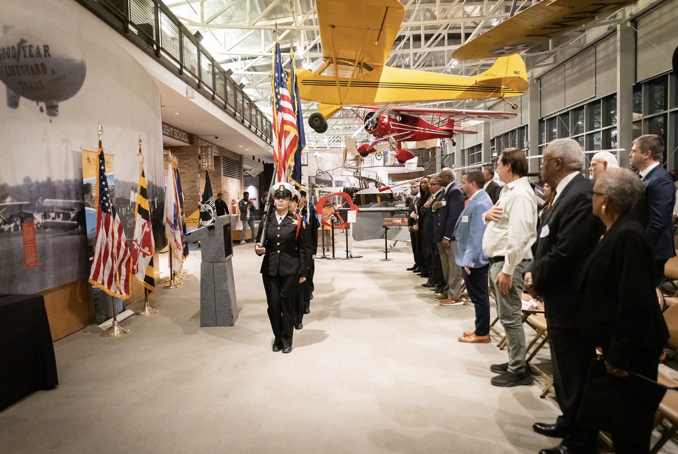 A group of people standing in a line inside a museum, with vintage aircraft hanging from the ceiling and flags displayed on the left side.