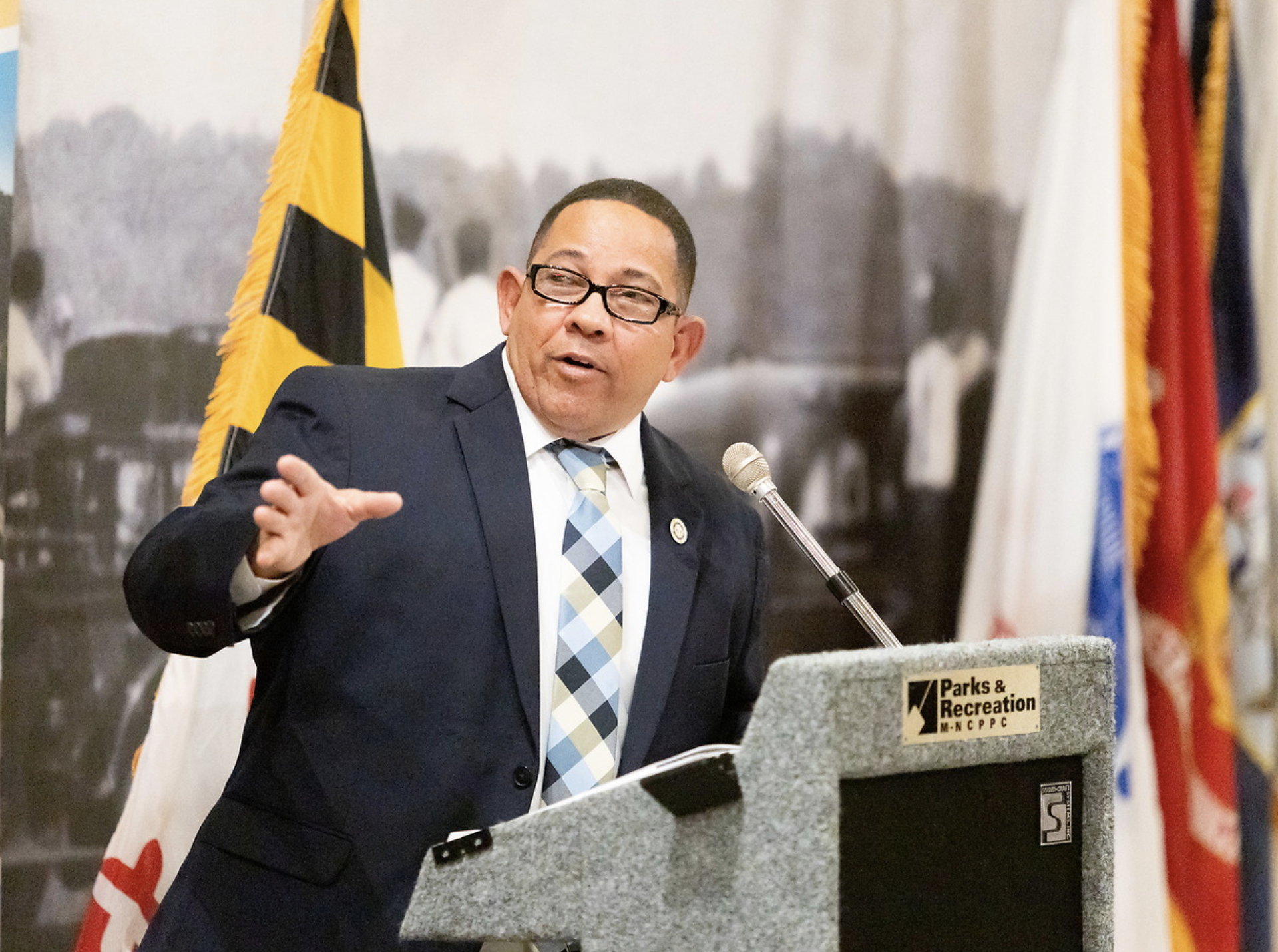 A man wearing glasses and a suit giving a speech at a podium with a microphone. Behind him are flags and a sign that reads 'Parks & Recreation'.