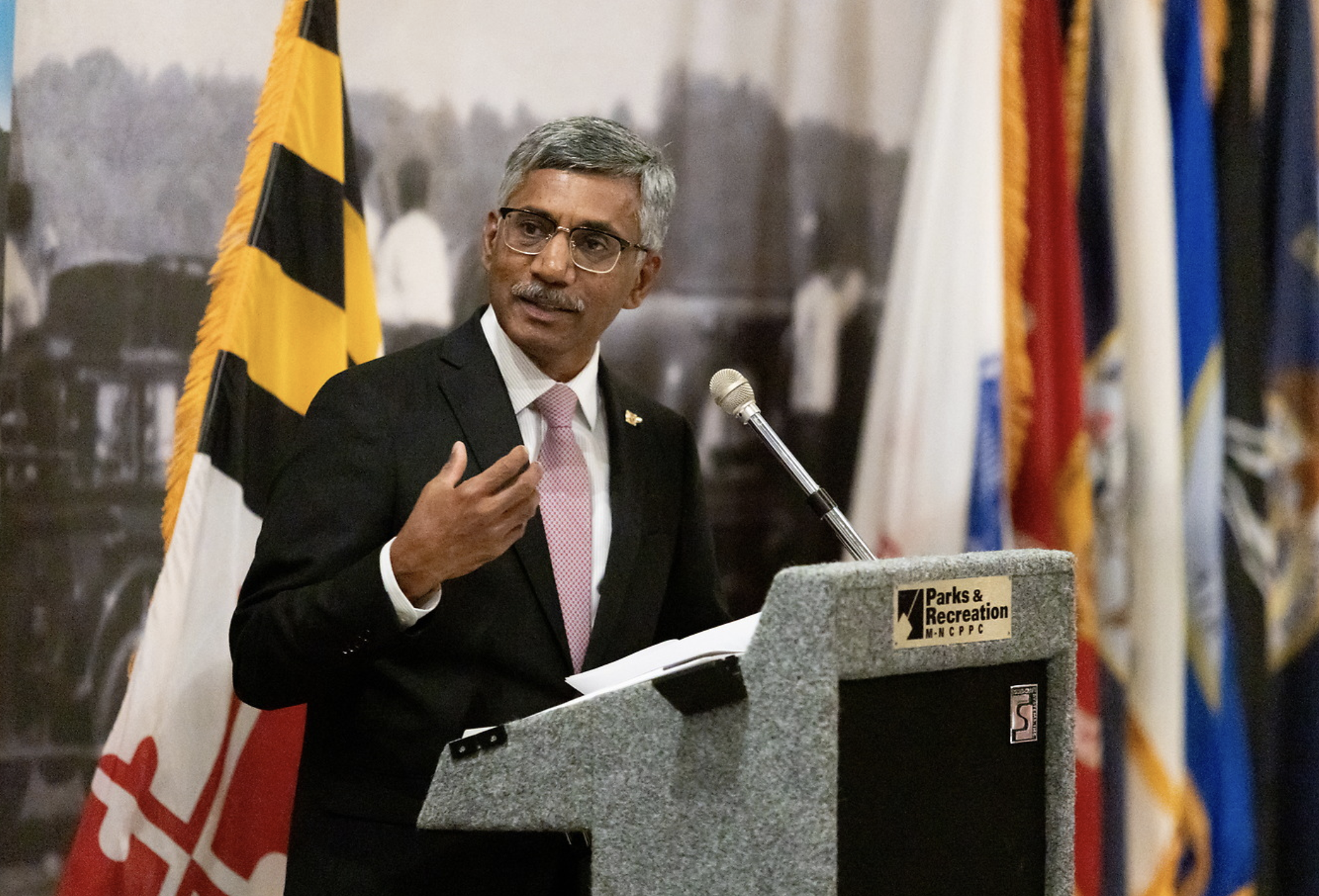 A man wearing glasses and a suit speaking at a podium during a public event, with flags in the background.
