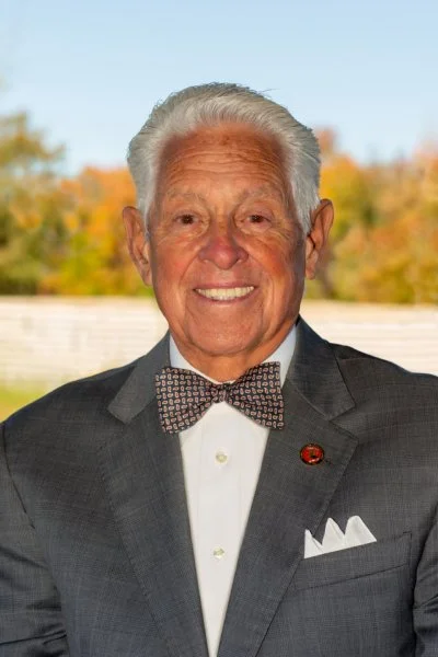 An elderly man with white hair, smiling, wearing a dark suit, a white shirt, a patterned bow tie, and a white pocket square, outdoors with autumn trees in the background.
