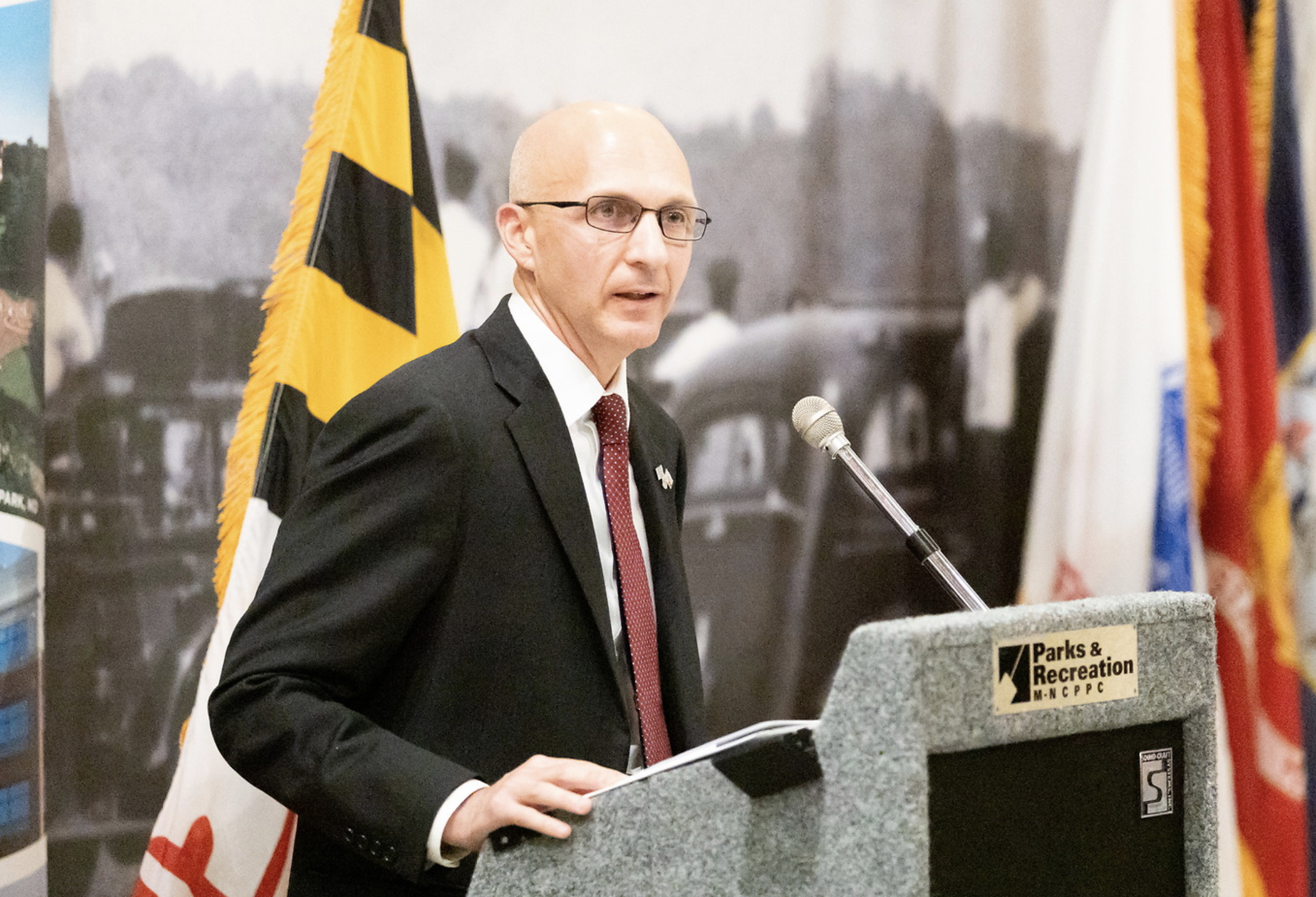 A man in a black suit and red tie speaking at a podium with a microphone, with a yellow and black flag, a partially visible American flag, and a historical photograph in the background.