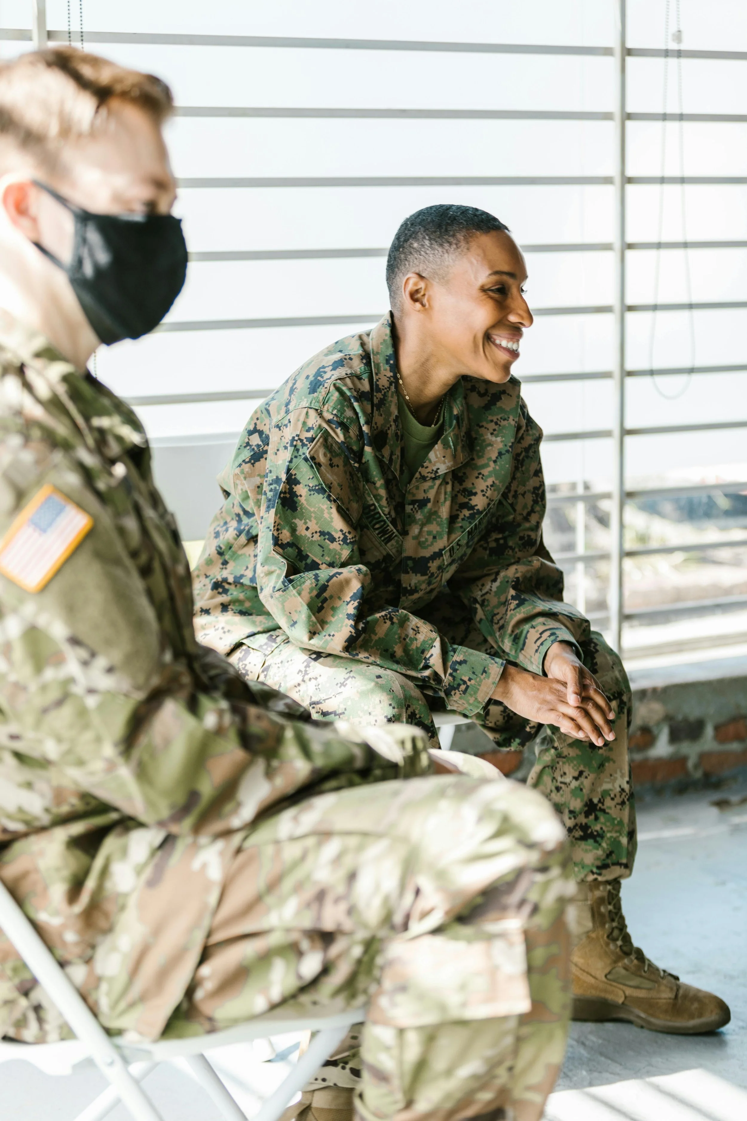 Two soldiers in camouflage uniforms sitting indoors near a window, smiling and engaged in conversation.