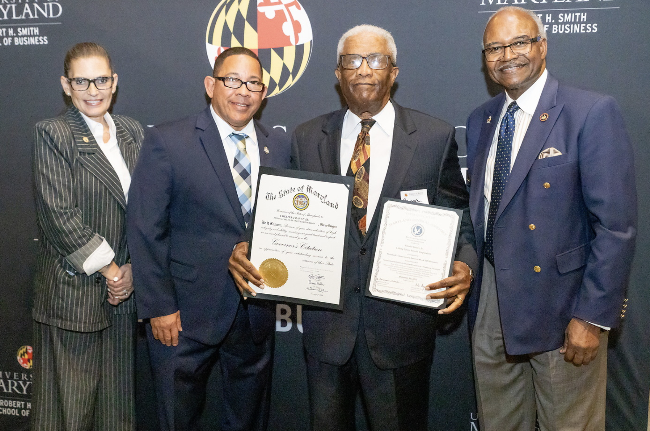 Four individuals in formal business attire standing together; two of them are holding certificates and smiling.