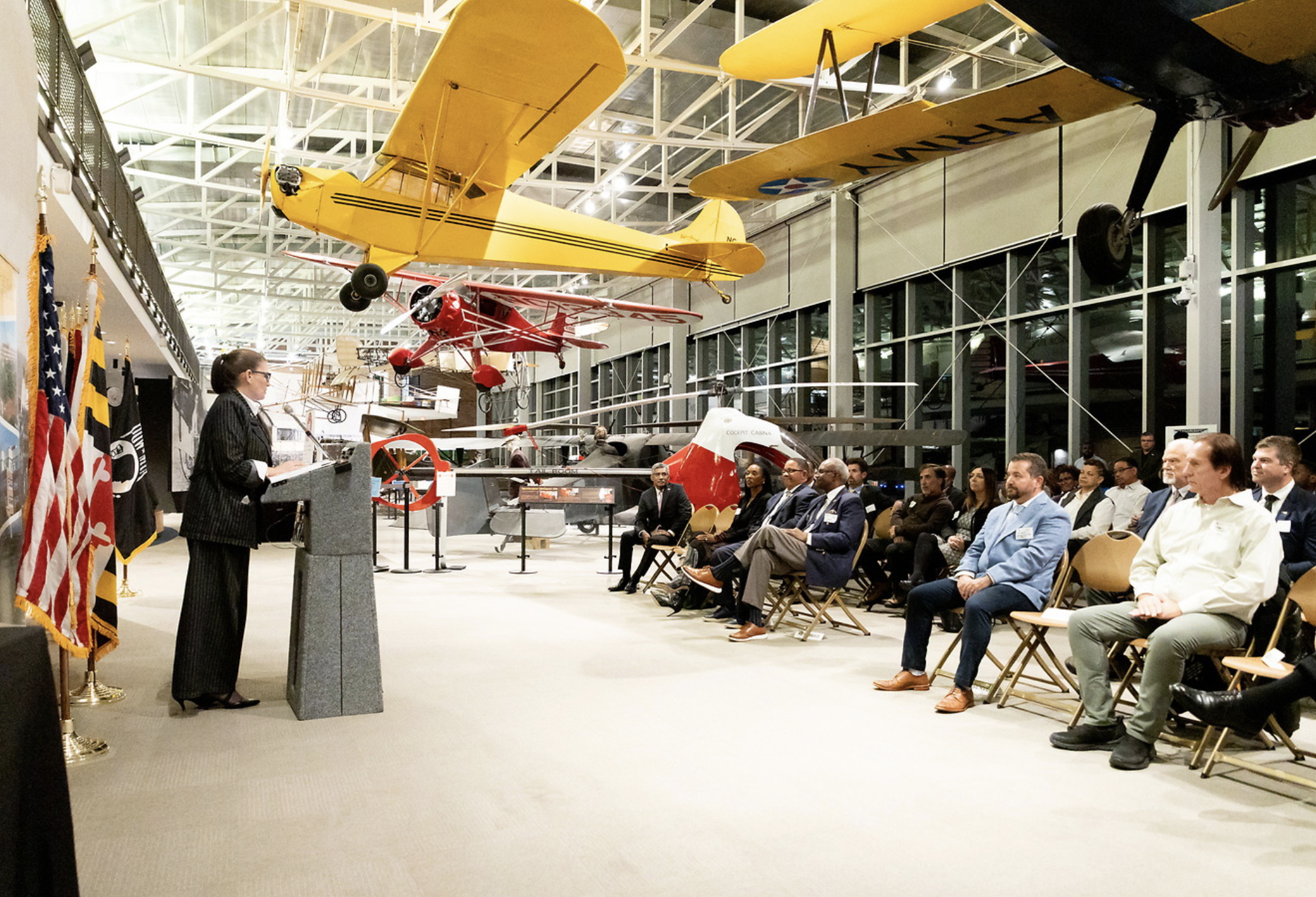 A woman in a black pinstripe suit speaking at a podium during a ceremony in an aviation museum, with vintage airplanes hanging from the ceiling and an audience seated in chairs.