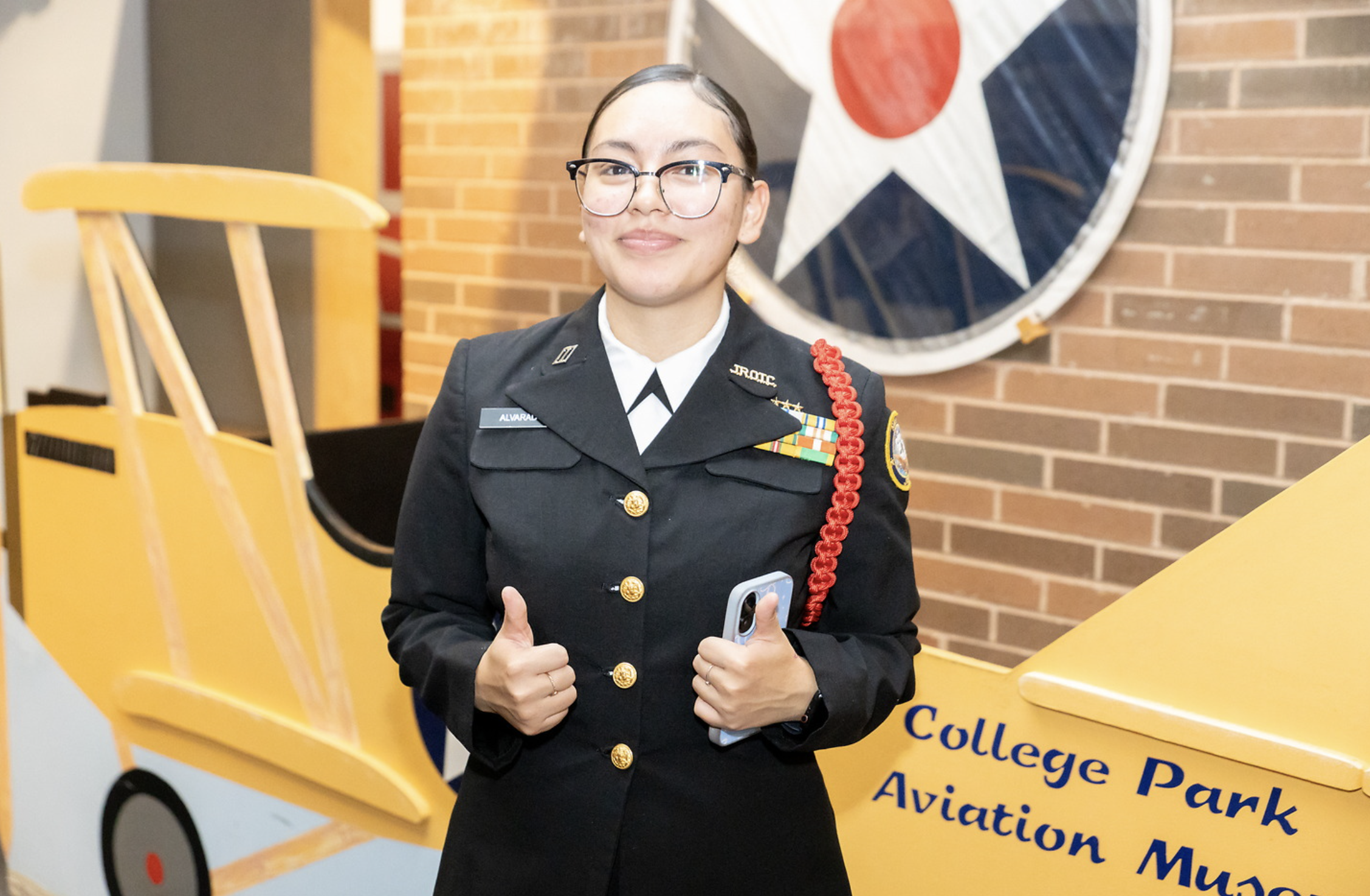 A young woman in a military uniform with glasses posing with a thumbs-up sign inside an airport or college park with a yellow cart that says 'College Park Aviation Museum'. She is standing in front of a brick wall and a large display of an airplane p