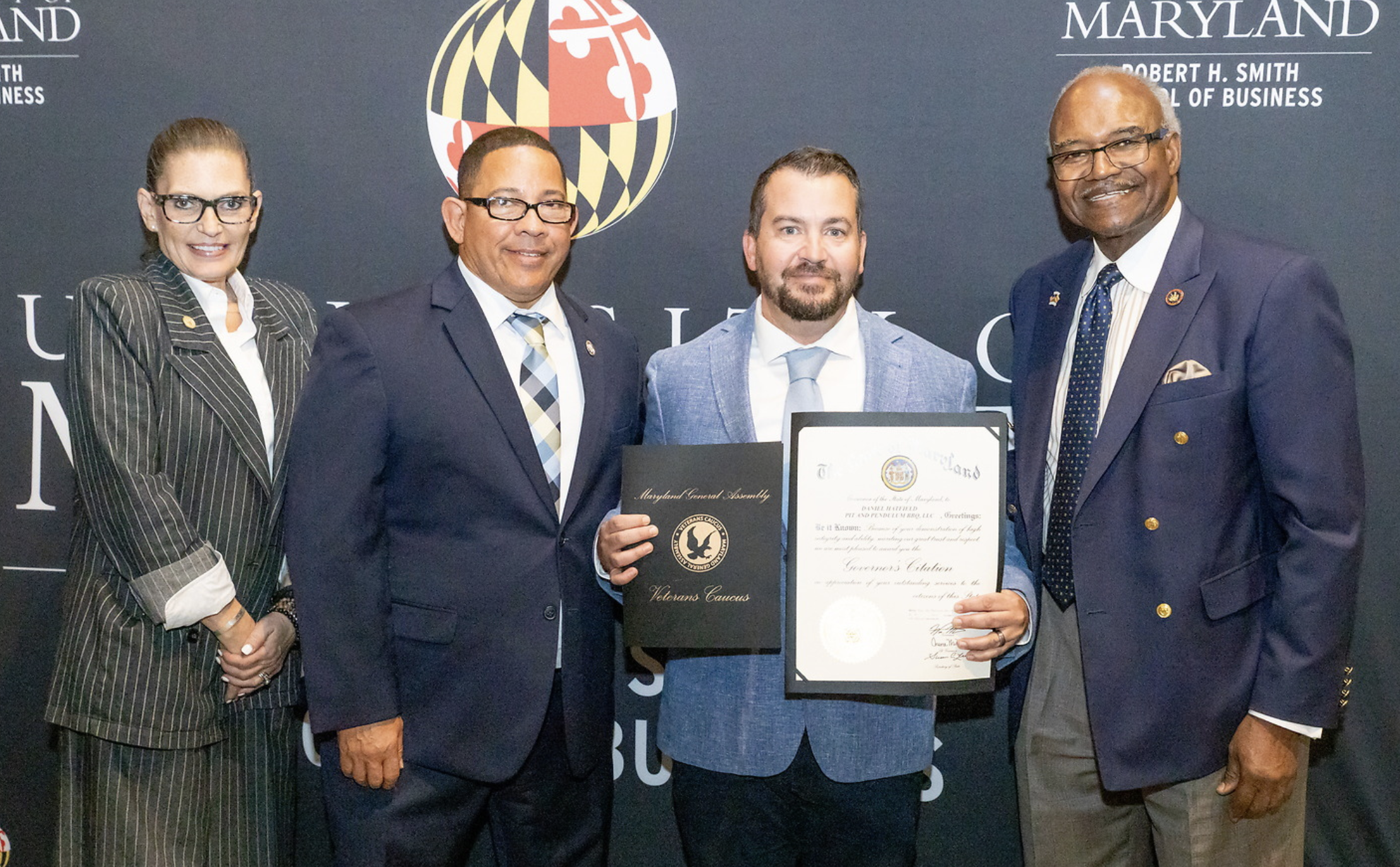 Four individuals standing together at a formal event, with a young man in the center holding a framed document and certificate, all dressed in business attire, in front of a backdrop featuring Maryland state logos and text.