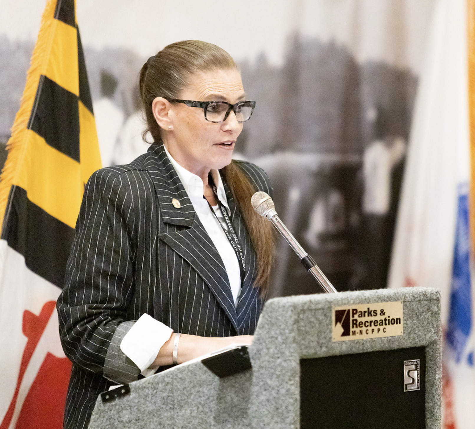 A woman with long brown hair, wearing glasses and a pinstripe blazer, stands at a podium speaking into a microphone. Behind her are flags and a blurred black-and-white photograph. The podium has a sign that reads 'Parks & Recreation' and 'M-NCPPC.'