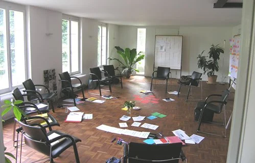 Ein leeres Meeting- oder Seminarraum mit Stühlen, Papier und Büchern auf dem Boden, große Fenster, Pflanzen, Whiteboard und Pinnwand.