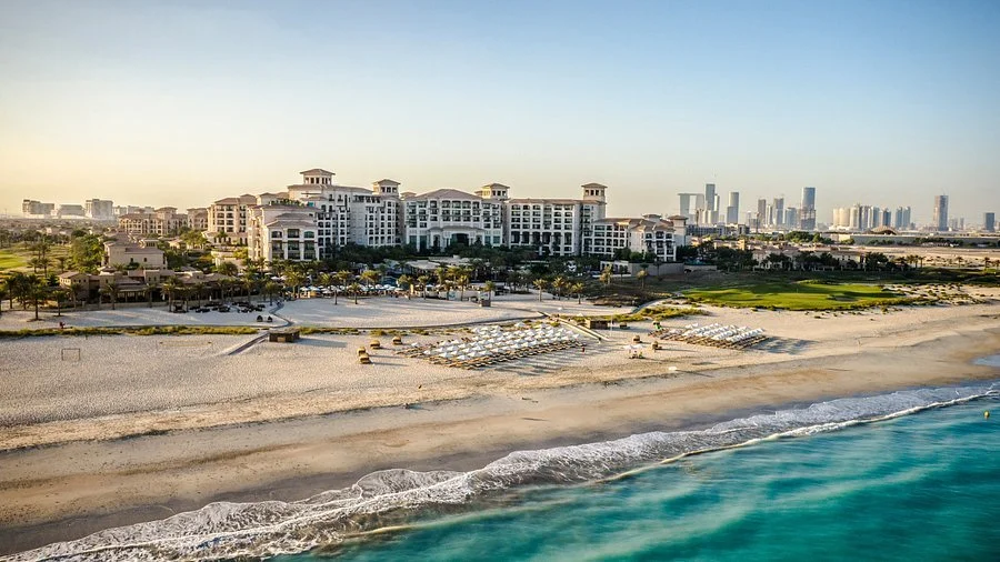 Aerial view of a beachfront hotel or resort with beach chairs, a beach, and city skyline in the background.