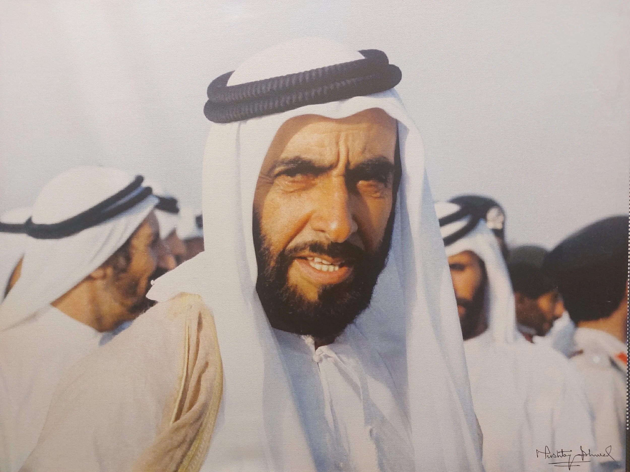A man with a beard in traditional Middle Eastern attire, including a white headscarf with black agal, standing among others dressed similarly.