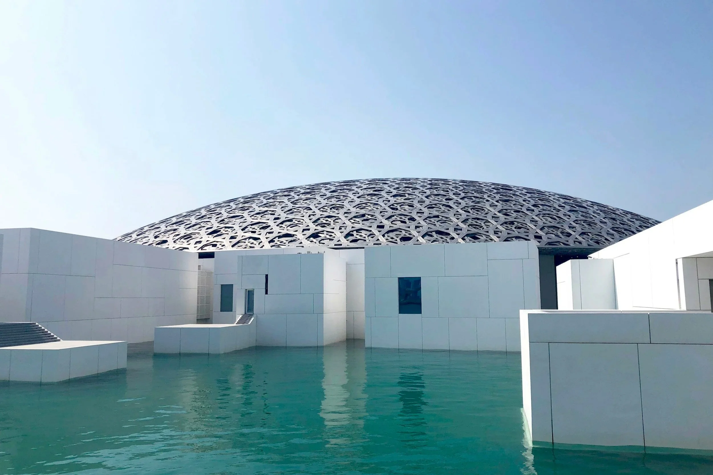 A modern building with a geometric patterned dome, white walls, and water in the foreground.