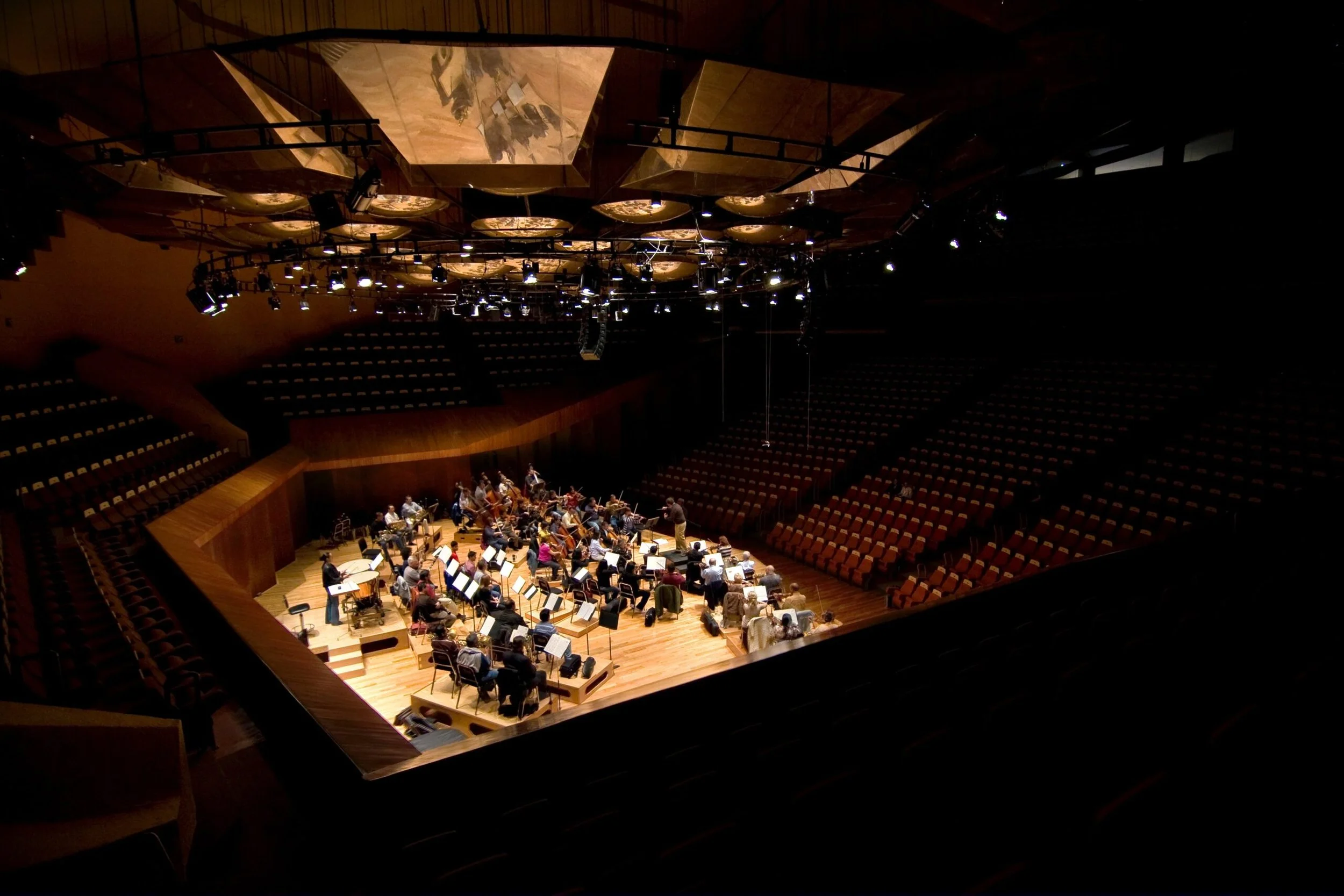 Orchestra rehearsing in a large concert hall with empty seats, viewed from an upper balcony.