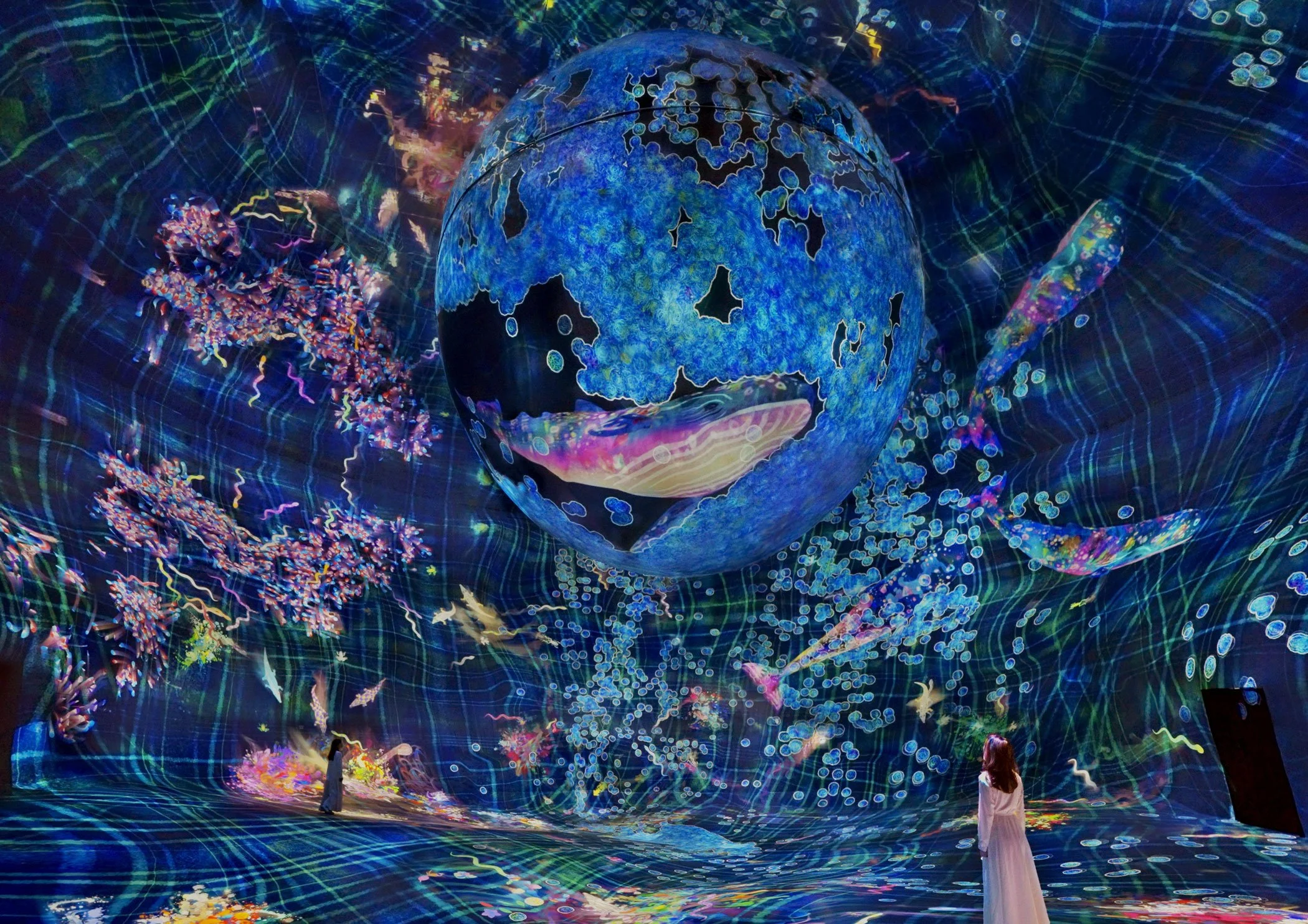 A large, spherical digital art installation depicts the Earth with colorful abstract patterns and surrounding vibrant light designs; two women in long dresses observe the display.