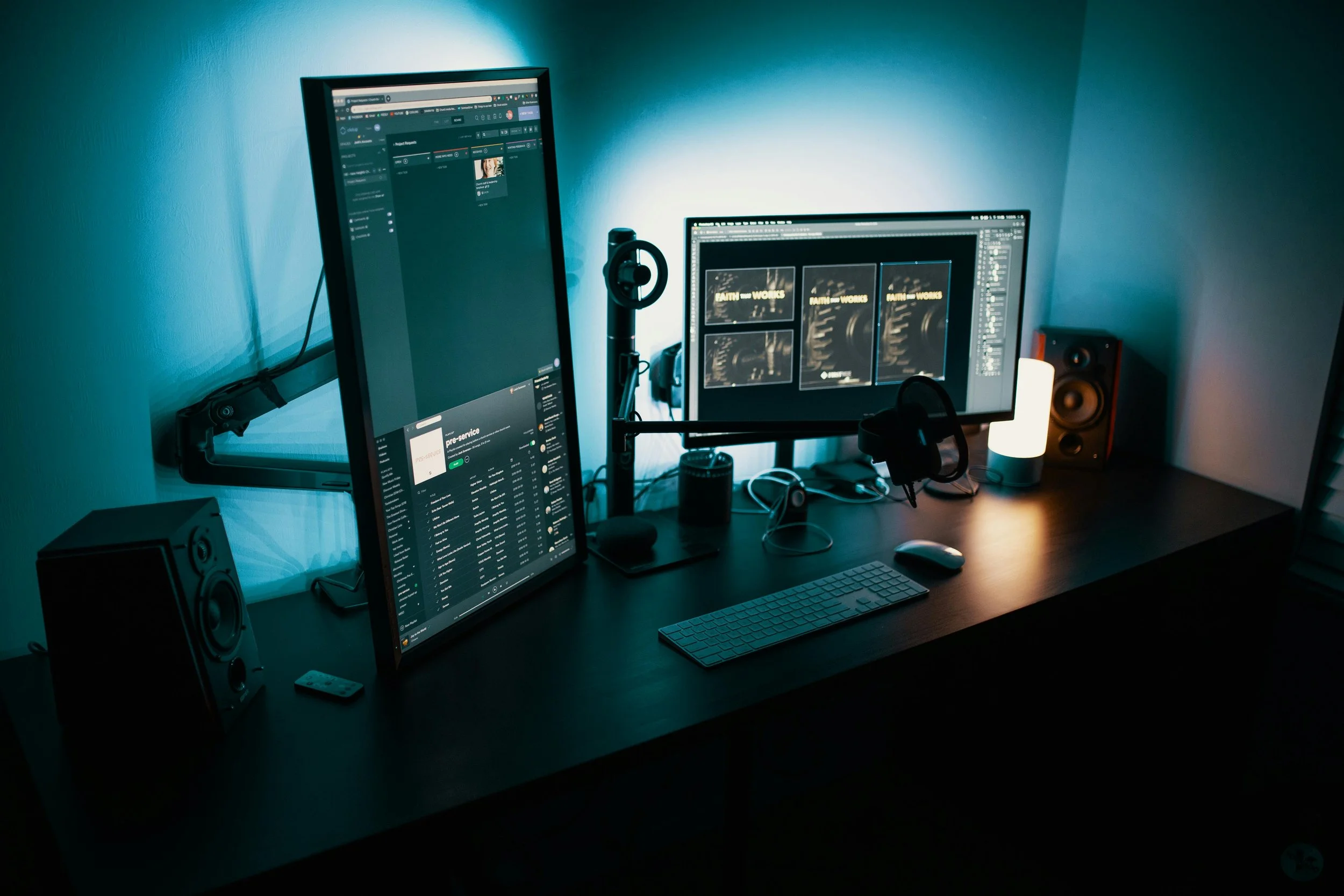 Modern computer workspace with dual monitors, speakers, a lamp, headphones, keyboard, mouse, and various electronic devices on a dark desk.
