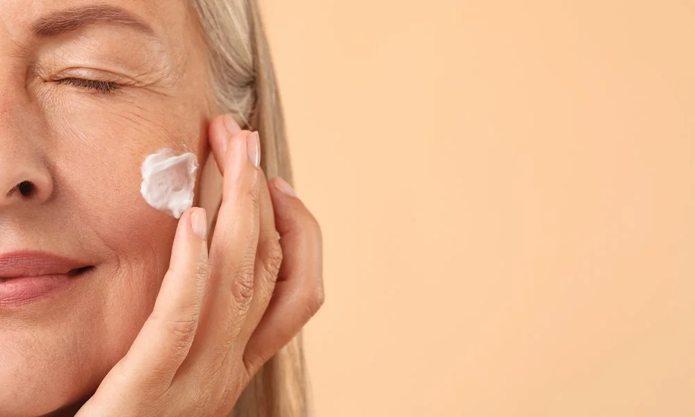 A woman applying cream or moisturizer to her face with her hand, smiling with her eyes closed.