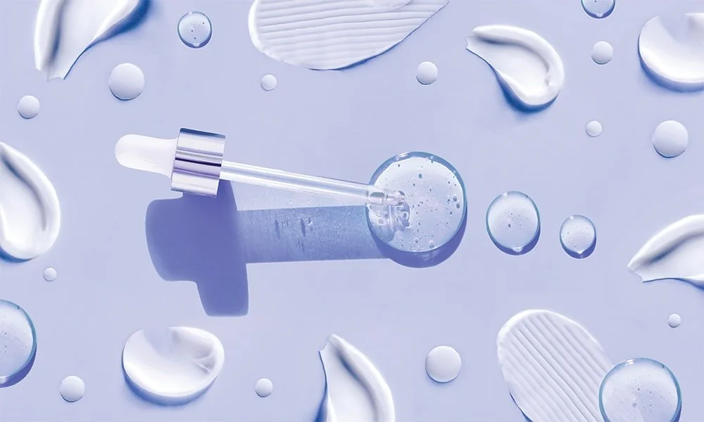A skincare serum dropper lying on a light blue surface with splashes and droplets of cream or lotion surrounding it.
