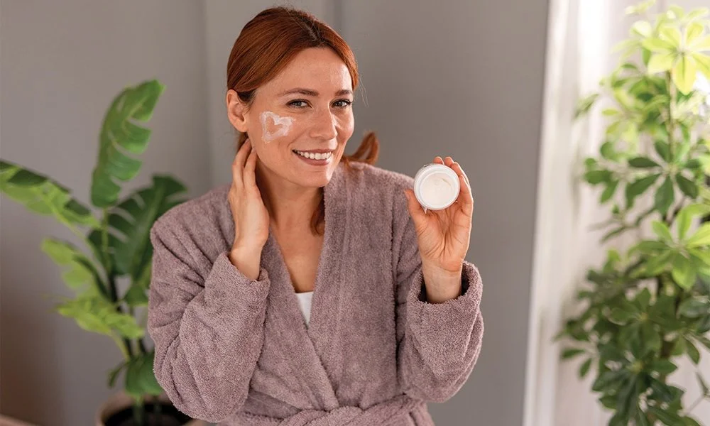 A woman with red hair smiling and holding an open jar of face cream, with some cream on her cheek.