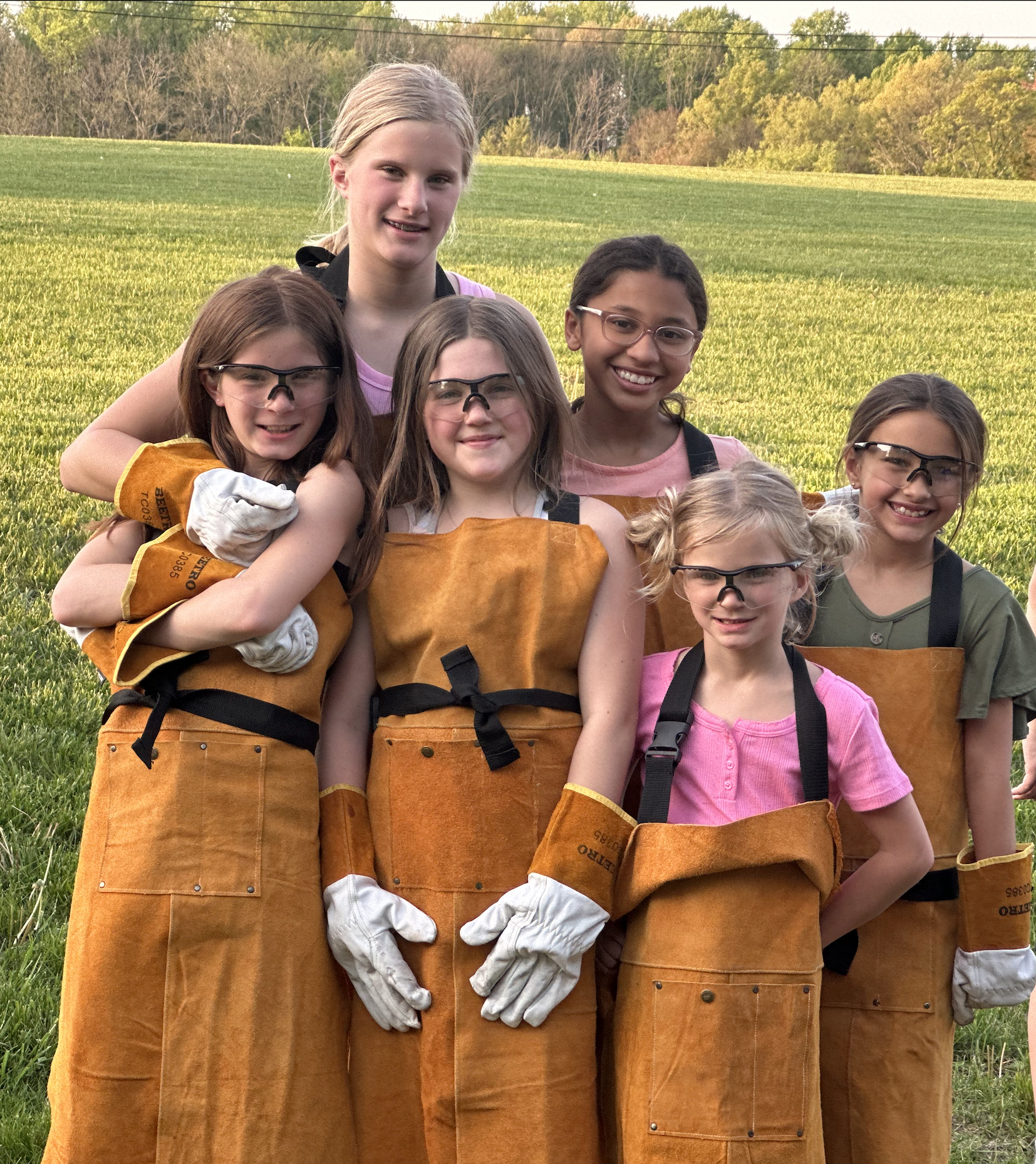Group of seven young girls wearing protective goggles, orange work aprons, and gloves standing outdoors on a grassy field with trees and a clear sky in the background.