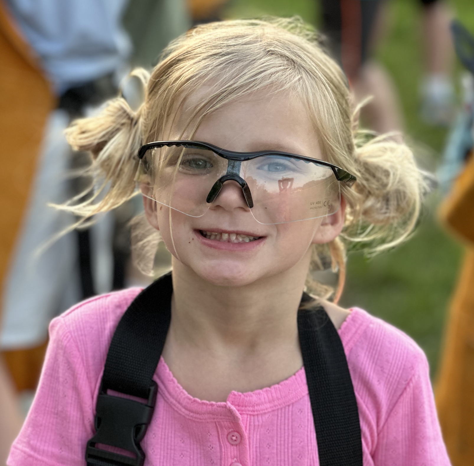 A young girl with blonde hair styled in two messy pigtails, wearing safety glasses, a pink top, and a black strap, smiling outdoors.