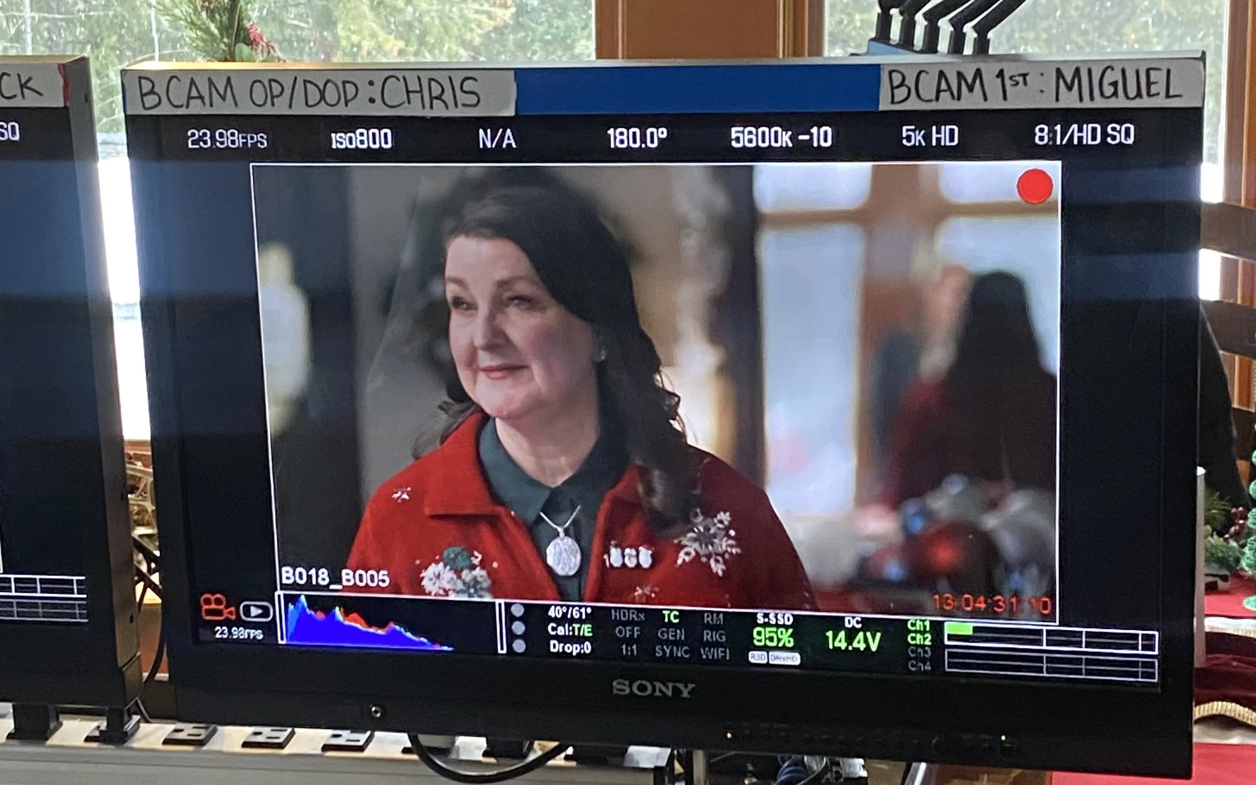 A woman on a film or video set being filmed with a Sony monitor displaying her on-screen. She is wearing a red Christmas-themed sweater with snowflakes and holly, and a necklace with a pendant. The background is blurred, showing an indoor setting with other people.