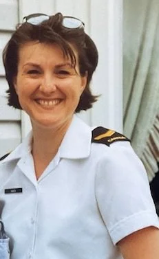 A woman smiling in a white uniform with a shoulder epaulet and a name tag, standing in front of a window with curtains.