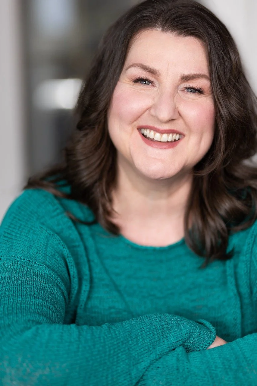 A woman smiling with dark brown, wavy hair wearing a teal sweater, sitting indoors.
