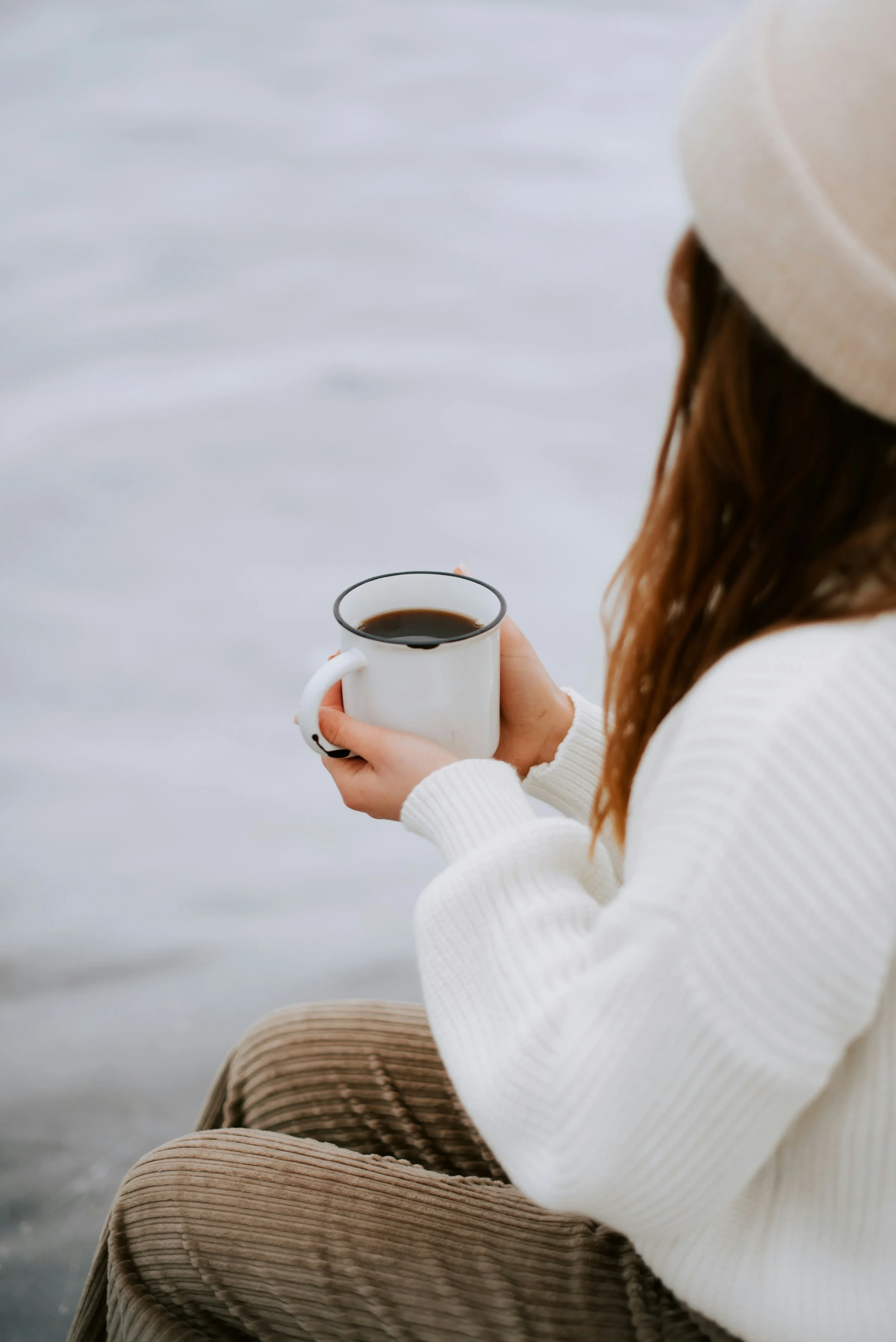 Person wearing a white knit sweater and beige corduroy pants, sitting by the water holding a white mug filled with coffee, with a beige hat on their head and brown hair.