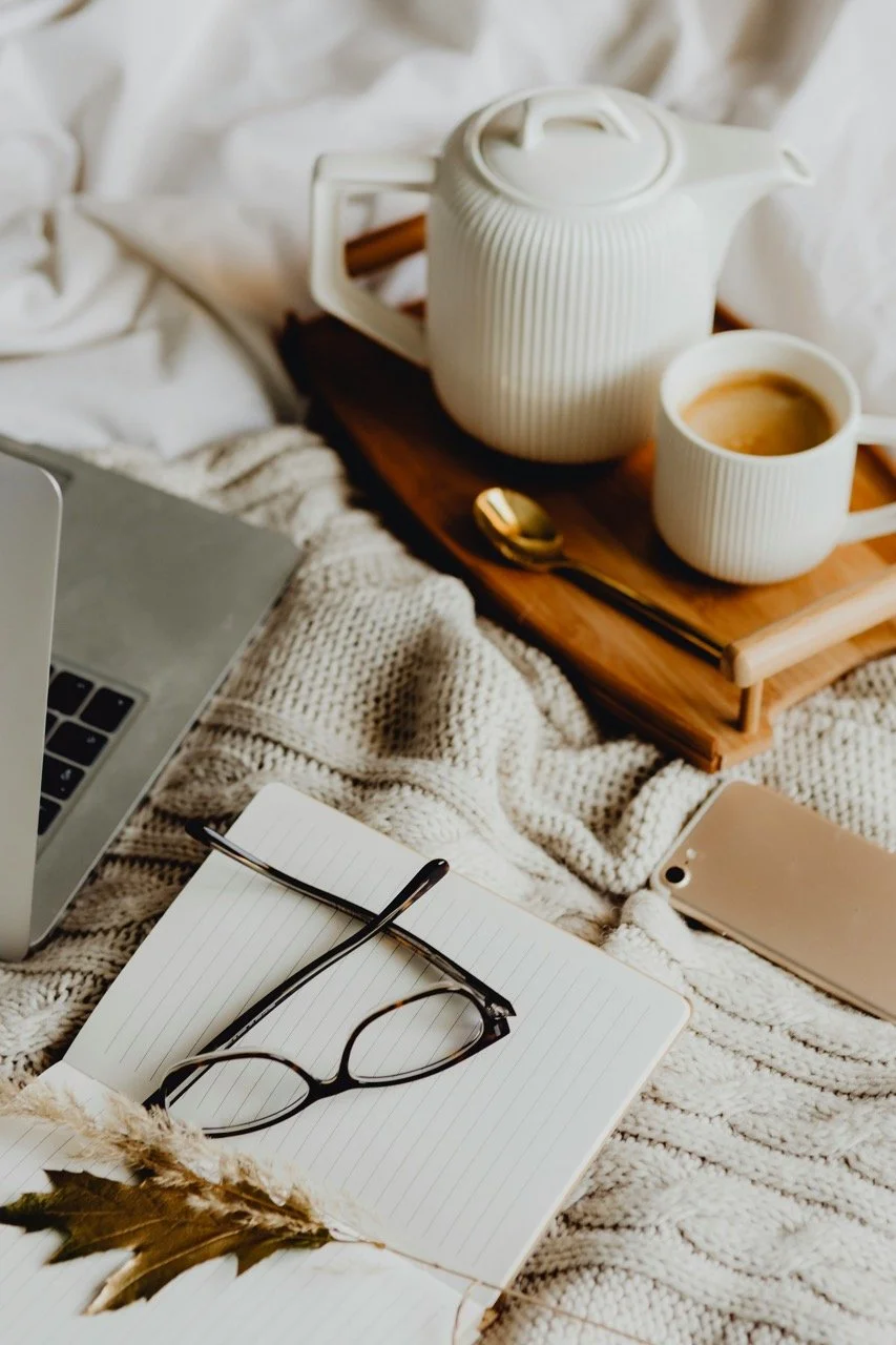 A cozy scene with a laptop, notebook with eyeglasses, a gold pen, a gold-colored smartphone, and a wooden tray with a white textured teapot and mug of coffee on a bed with white knitted blankets.