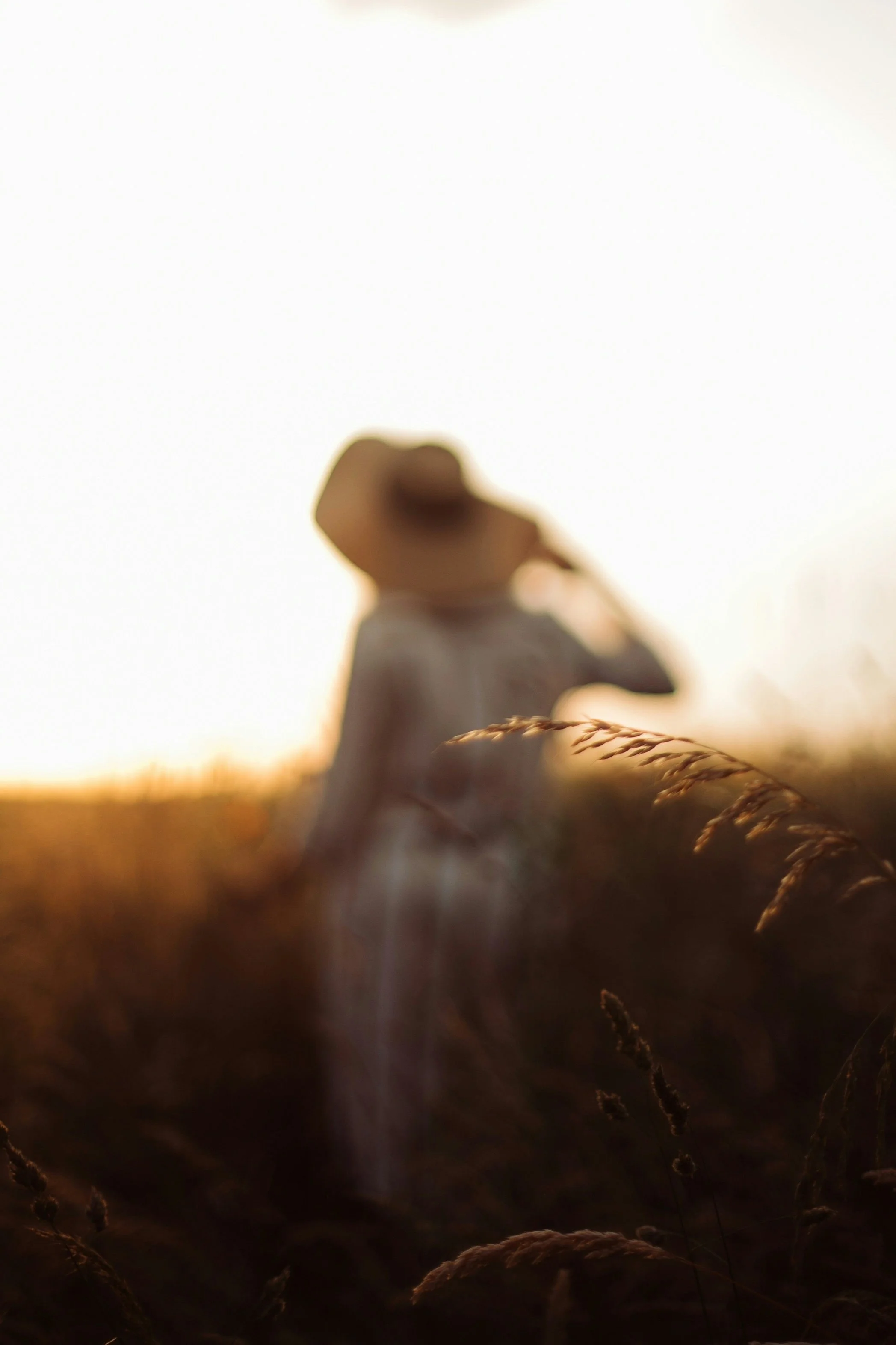 A blurred person wearing a hat standing in a field of tall grass during sunset.