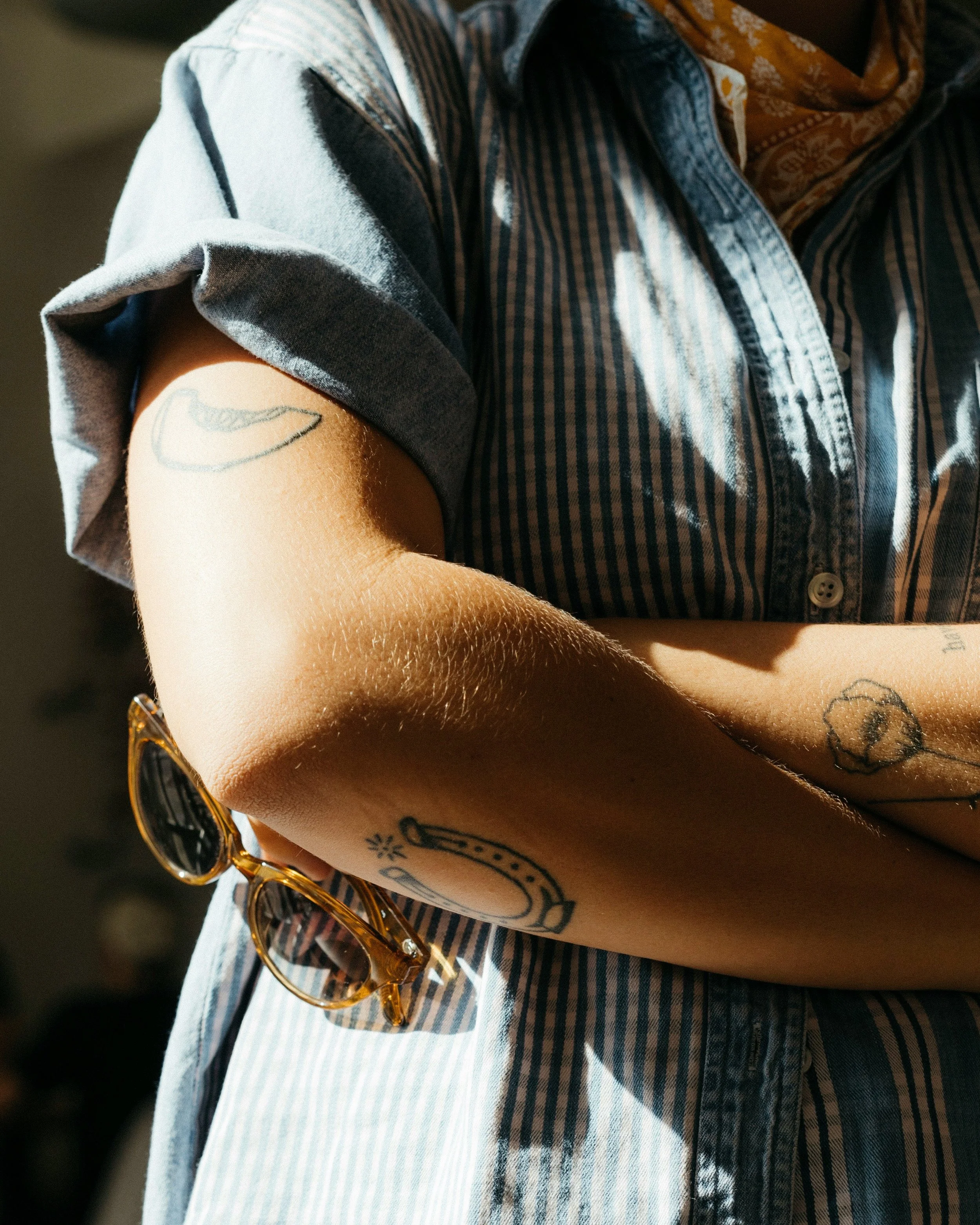 Close-up of a person with sleeve rolled up, showing tattoos on their arm, holding sunglasses, wearing a striped shirt and a bandana around their neck.