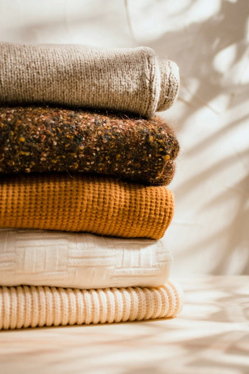 Stack of folded sweaters in neutral tones on a light surface.