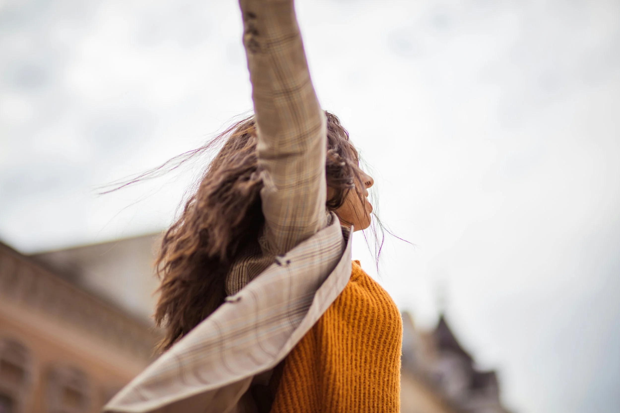 Woman with brown wavy hair wearing a yellow sweater and beige jacket, raising her arm outdoors, with a cloudy sky and historic buildings in the background.