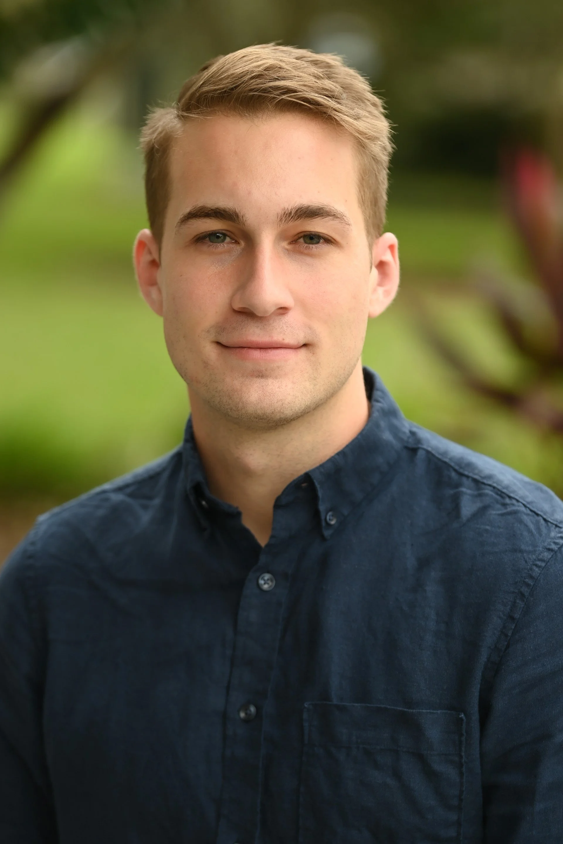 A young man with blonde hair wearing a dark blue button-up shirt standing outdoors with a blurred green background.