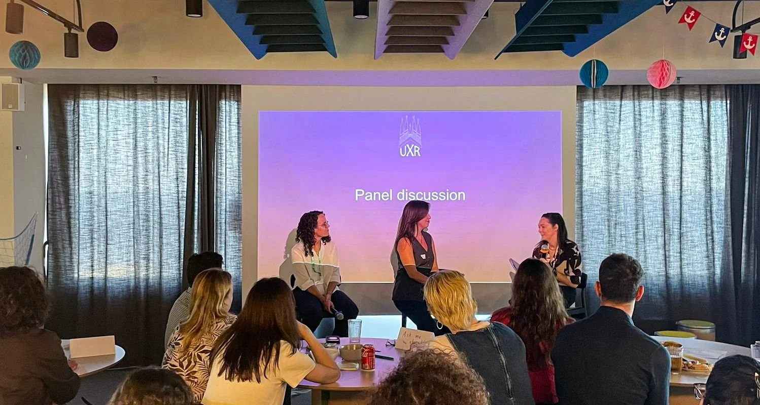 Panel discussion at a UX conference with three women on stage, audience seated, with a purple projection screen displaying 'UX', 'Panel discussion'.