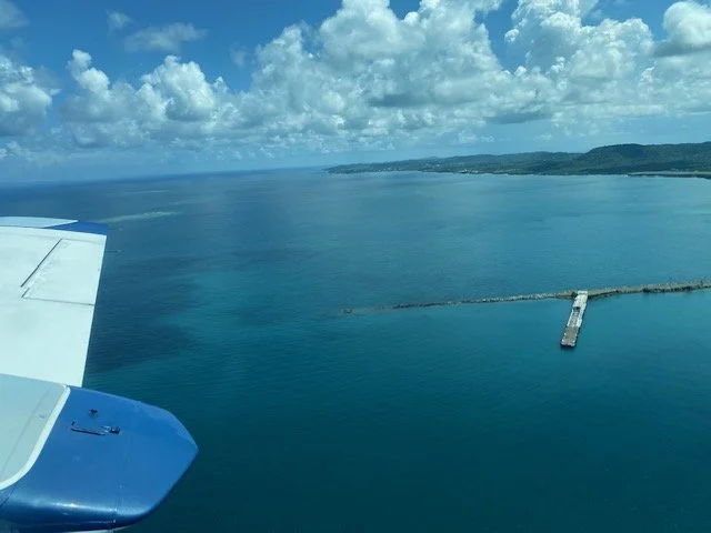 Aerial view of a coastline with a pier extending into the water, taken from an airplane window showing part of the wing and engine.