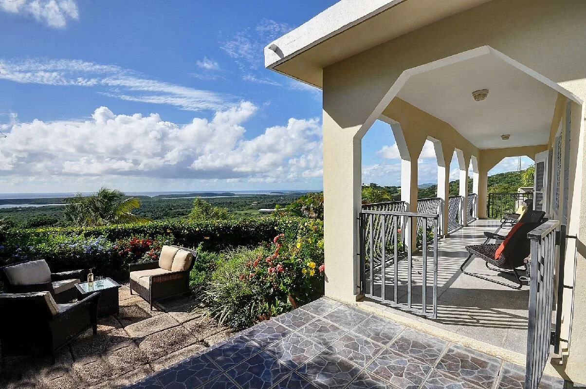 View from a balcony showing outdoor seating and a lush green landscape with views of the ocean.